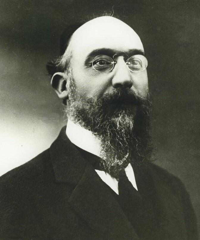 Francúzsky skladateľ a klavirista Erik Satie na historickej čiernobielej fotografii. Má okuliare, dlhú bradu a formálny oblek.