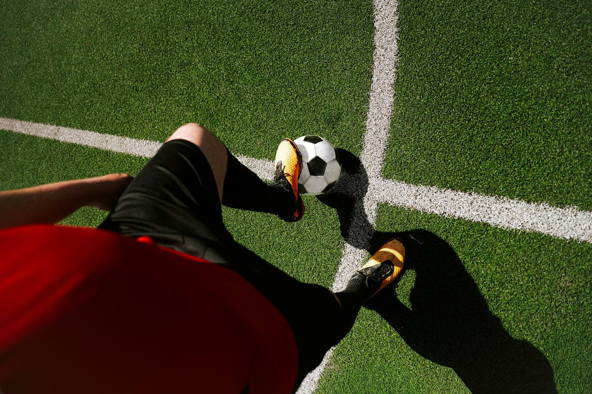 Futbalista stojí na ihrisku s nohou položenou na futbalovej lopte pri bielej čiare, pripravený na začiatok hry.