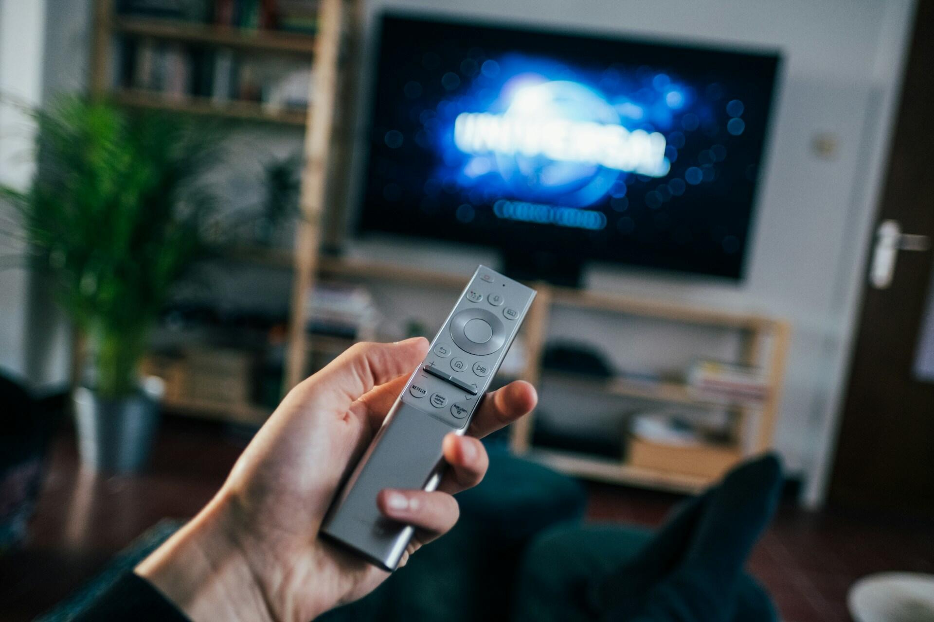 Ruka s diaľkovým ovládačom pred televízorom počas sledovania seriálu.