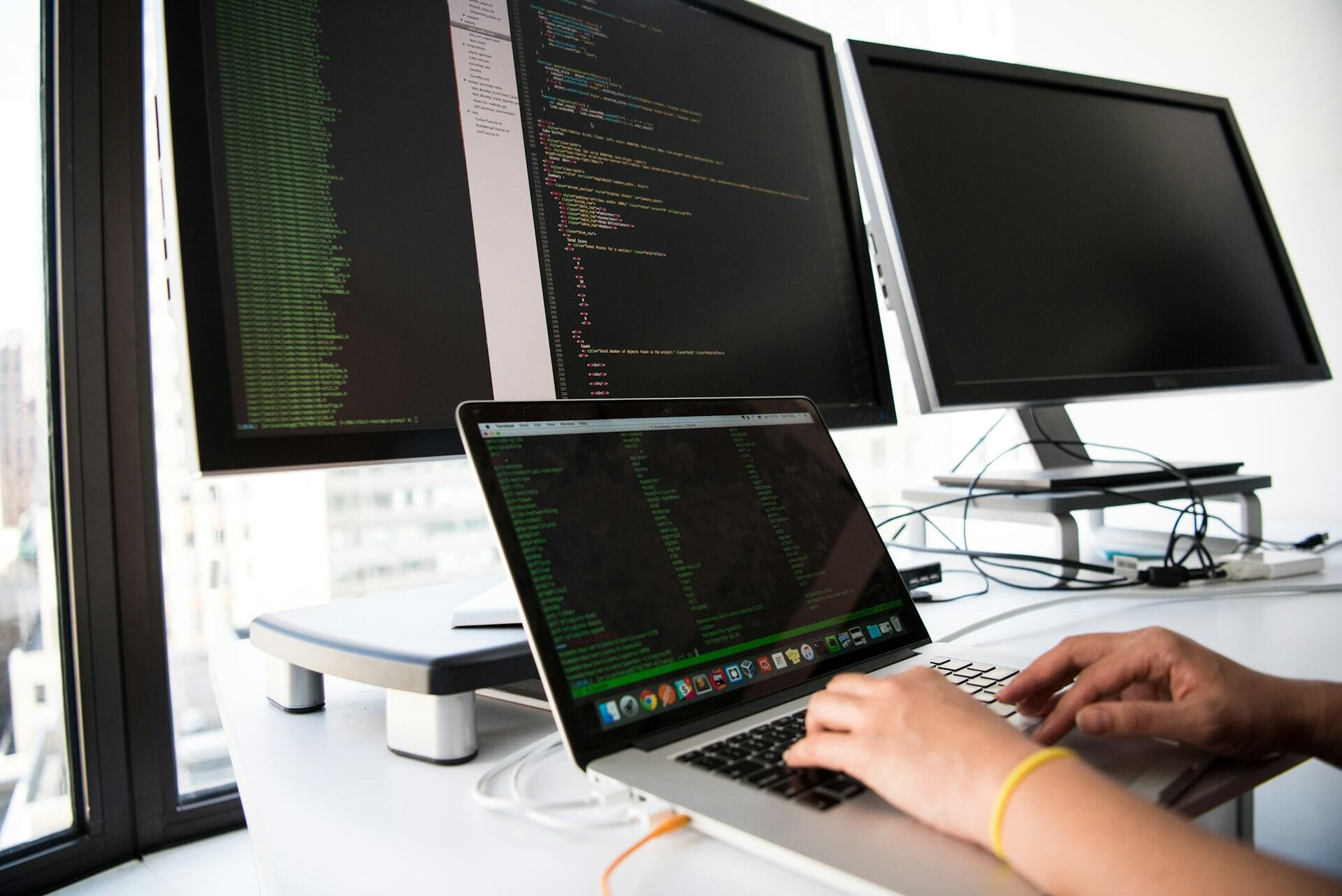 Programátor pracuje na notebooku s otvoreným zdrojovým kódom a viacerými monitormi.