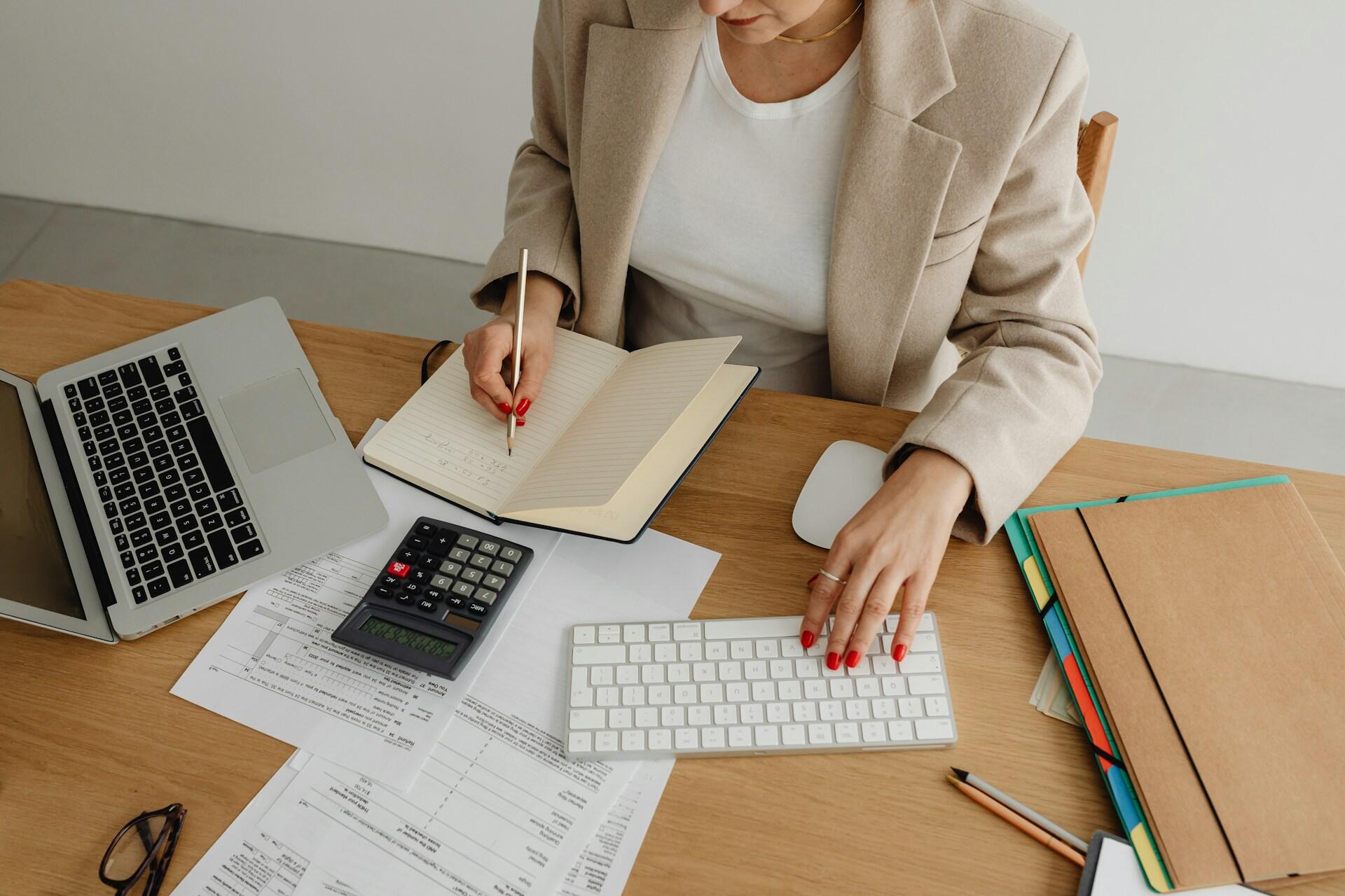 Účtovníčka pracuje s finančnými dokumentmi, kalkulačkou a zapisuje si poznámky pri počítači.