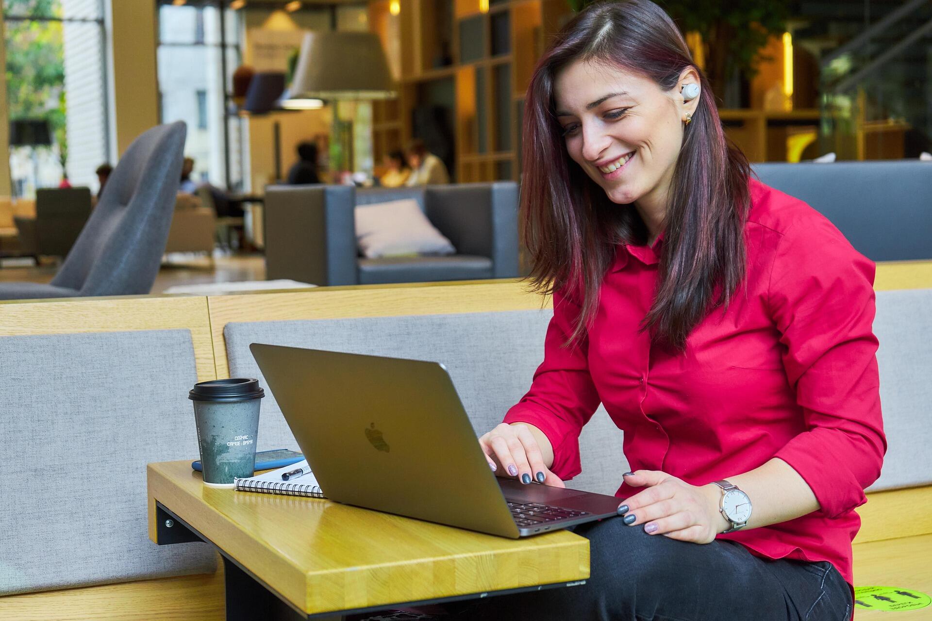 Geniş ve modern bir kafe veya çalışma alanında, ahşap bir masa üzerinde duran MacBook marka bir dizüstü bilgisayarla çalışan, kırmızı gömlekli, gülümseyen genç bir kadın.