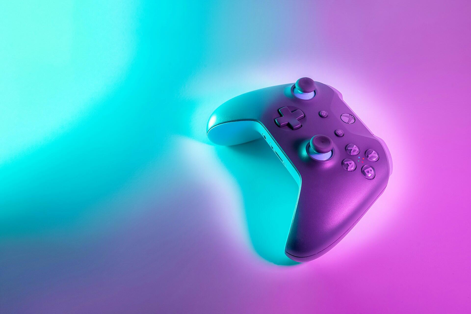 Turkuaz ve macenta neon ışıklarla aydınlatılmış, minimalist bir zemin üzerinde duran modern bir oyun konsolu kolu (gamepad).