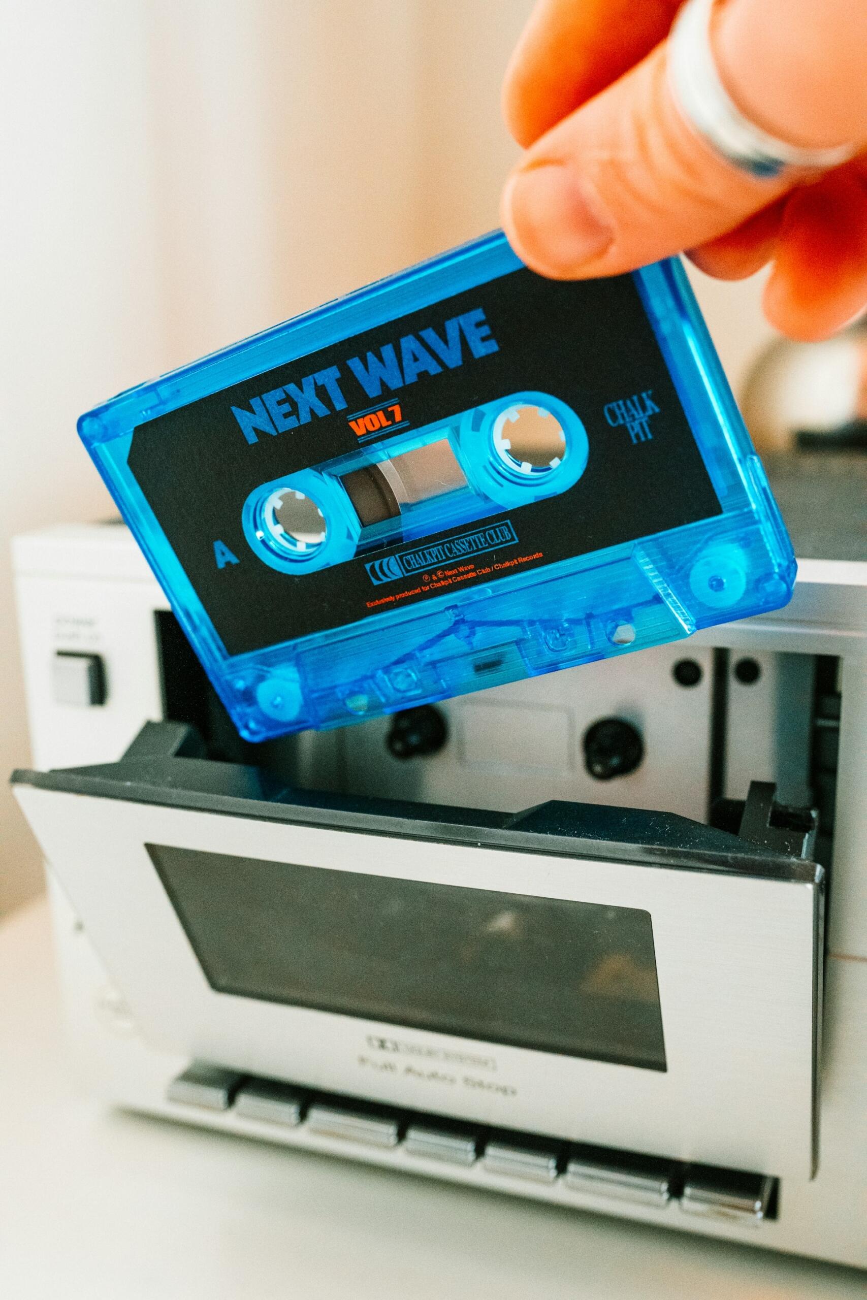 90'lar kaset çalarına mavi renkli, "Next Wave Vol. 7" etiketli kaseti takan el; kaset çaların kapağı açık.