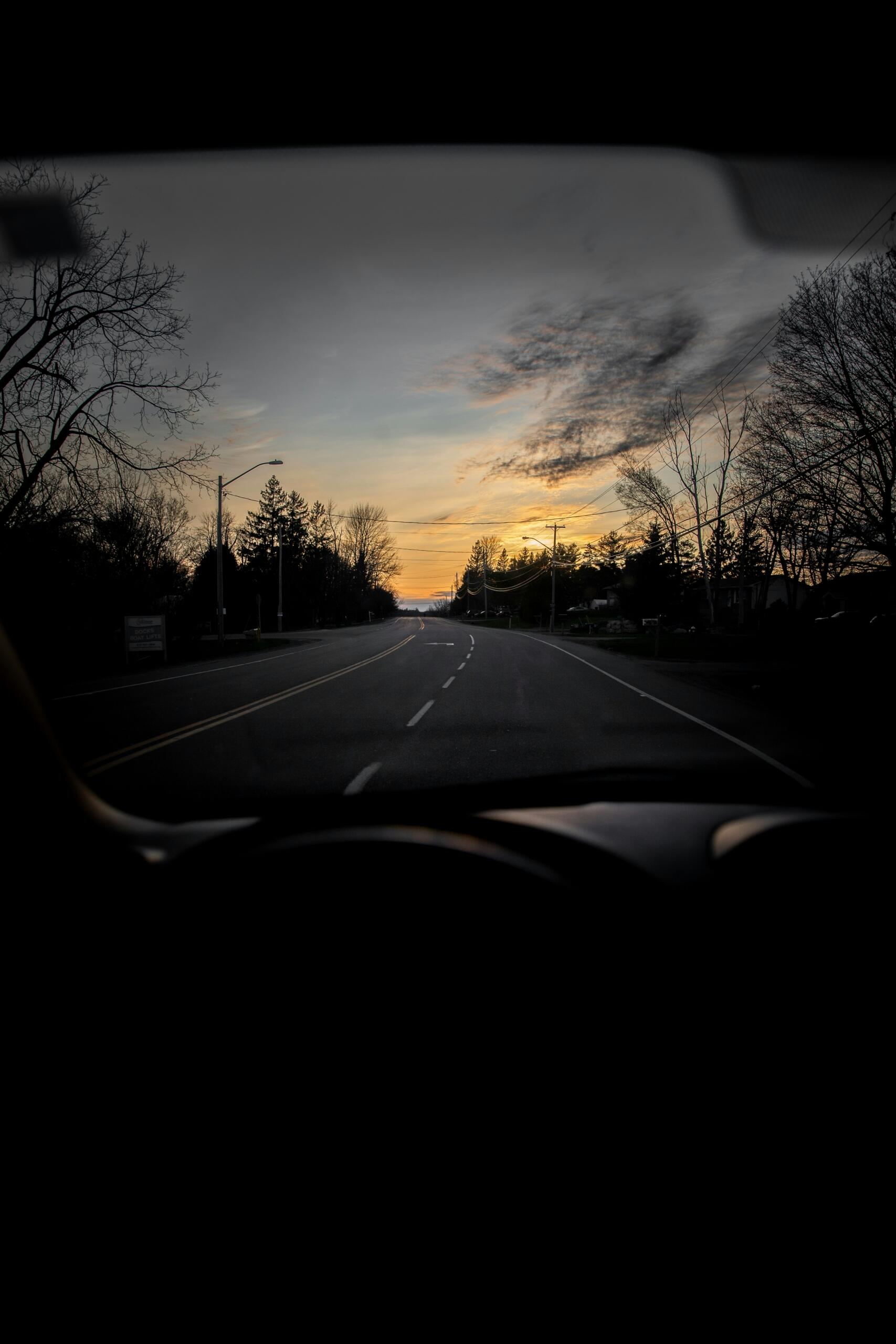 Bir otomobilin içinden ön cam aracılığıyla çekilen, gün batımının turuncu ve sarı renklerinin ufukta ağaçlar arasından süzüldüğü ıssız bir asfalt yol manzarası.