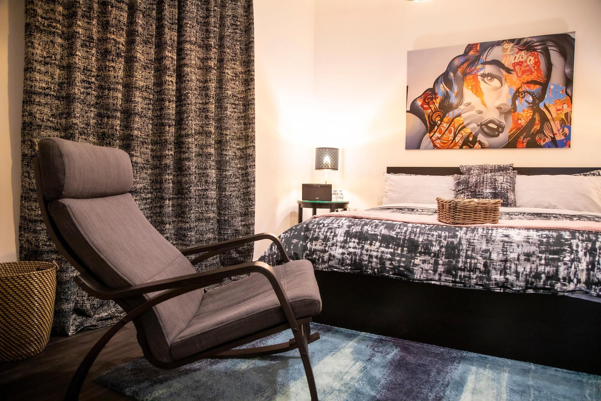 Modern yatak odası dekorasyonu: Gri Eames tarzı sallanan sandalye, pop-art duvar sanatı, desenli yatak örtüsü ve dokuma hasır sepet ile sıcak ve şık bir iç mekan.