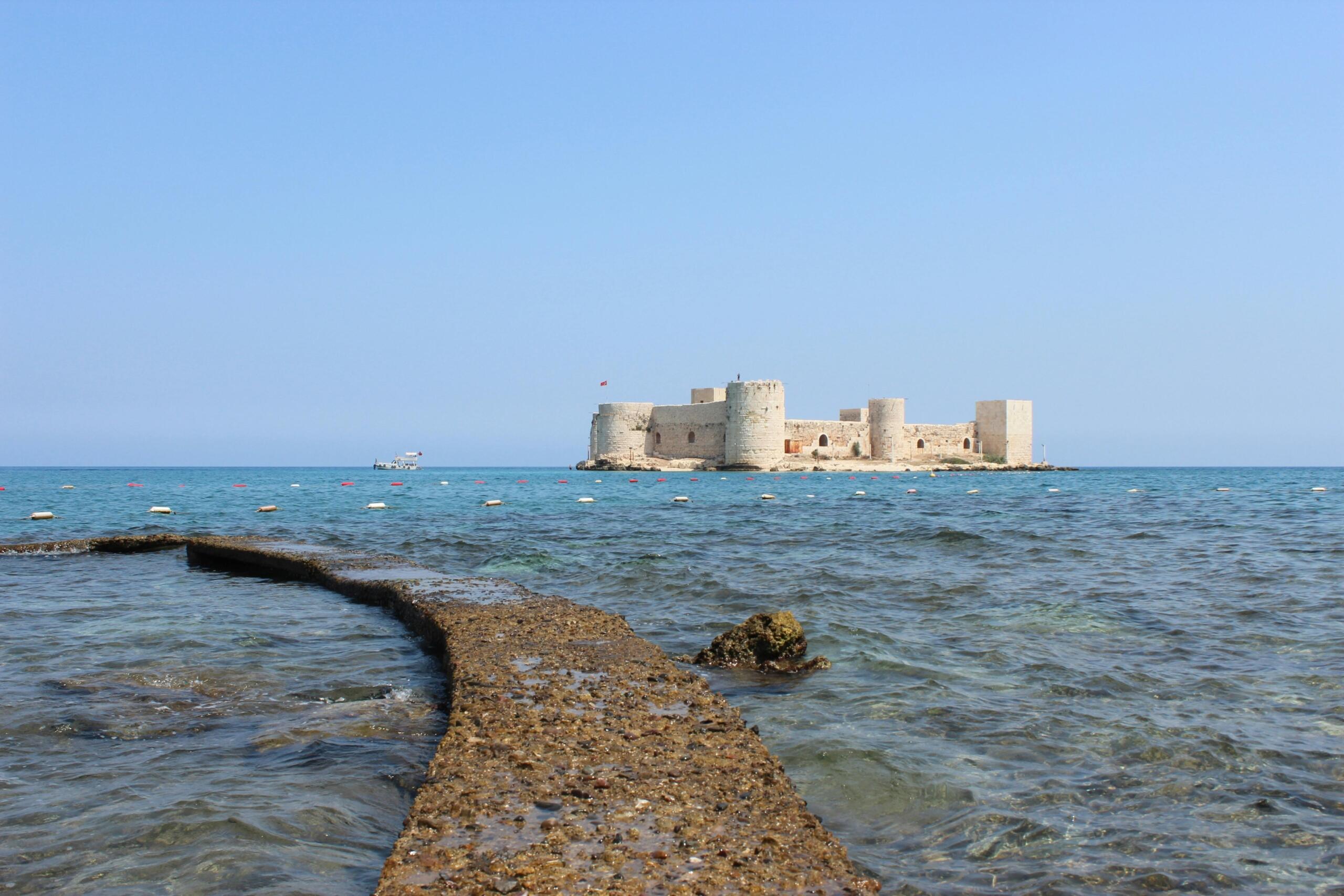 Masmavi denizin ortasında, ufukta yer alan Kızkalesi'nin, kıyıdan suya doğru uzanan taş bir yol perspektifinden çekilmiş geniş bir manzarası.