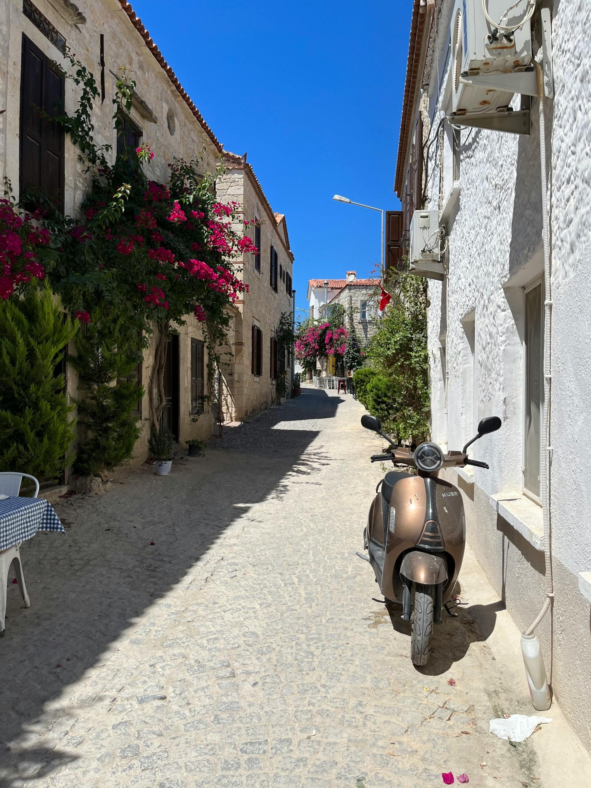 Geleneksel taş binaların ve canlı pembe begonvillerin süslediği dar bir sokakta, park edilmiş kahverengi bir motosikletin ön planda olduğu, güneşli ve huzurlu bir Alaçatı manzarası.