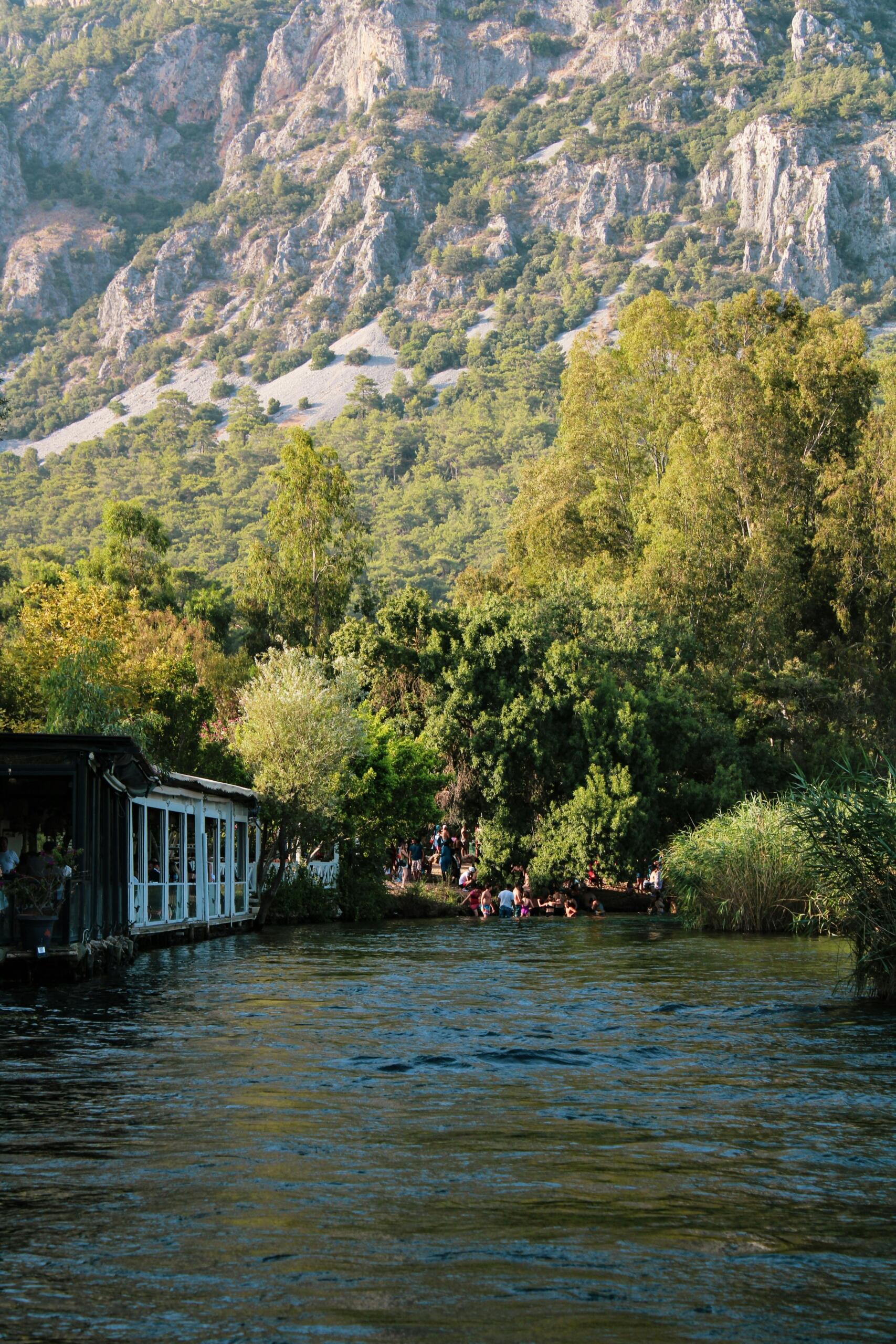 Sık ağaçlarla çevrili ve arkasında heybetli kayalık dağların yükseldiği nehir kıyısında, insanların suya girdiği ve sol tarafta beyaz çerçeveli pencereleri olan ahşap bir yapının bulunduğu Akyaka manzarası.