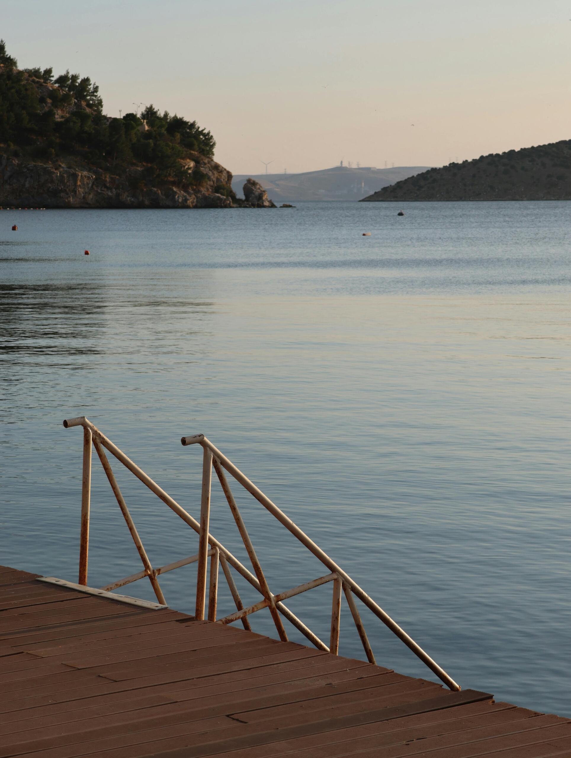 Ahşap bir iskelenin üzerinden denize inen metal merdivenlerin görüldüğü, arka planda ise tepelerin çevrelediği sakin ve durgun bir koyun gün batımı ışığındaki görünümü. Erdek
