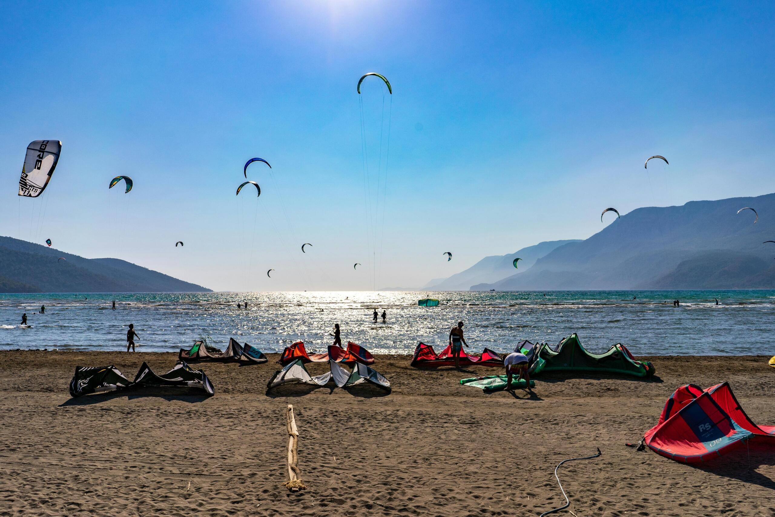 Dağlarla çevrili Akyaka Plajı'nın çok sayıda kiteboard sporcusunun havada süzülen renkli uçurtmalarıyla denizin ve kumsalın tadını çıkardığı bir manzara.