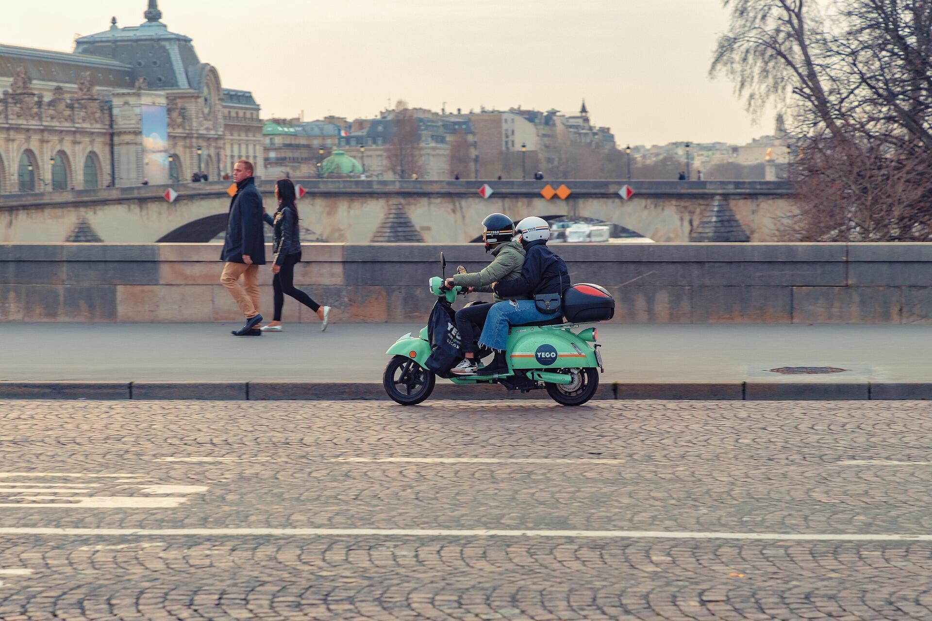 Şehir içinde ilerleyen iki kişilik YEGO marka yeşil scooter. Arka planda tarihi bir köprü ve şehir manzarası yer alıyor. Scooter, 125 cc’lik motoruyla hafif, hızlı ve ekonomik ulaşım sağlıyor.