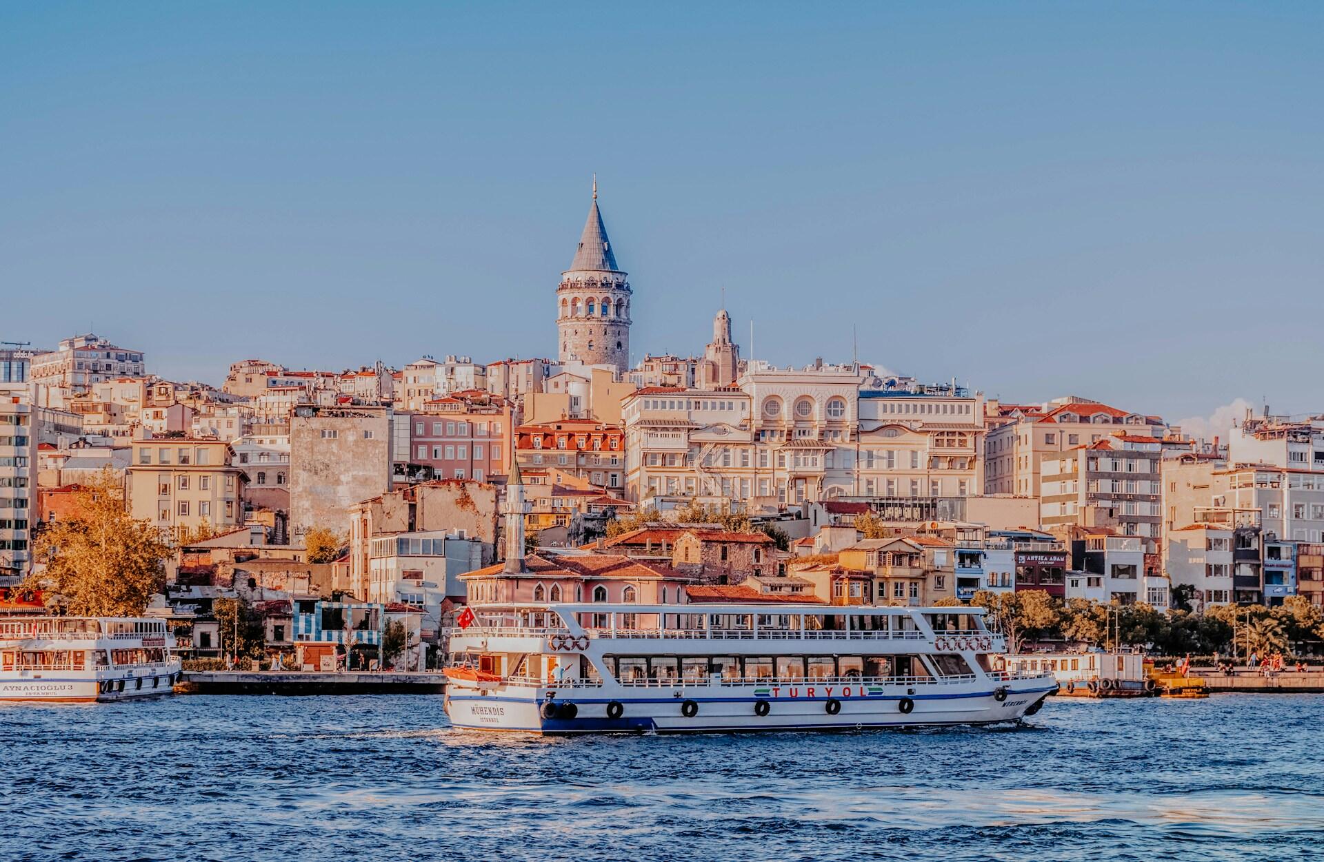 İstanbul’un Galata Kulesi manzarası, Haliç üzerinde yol alan bir vapur eşliğinde, gün batımında şehir silüetiyle birlikte.