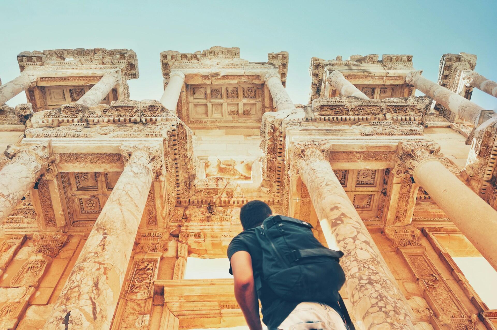 Sırt çantalı bir gezgin, Efes Antik Kenti’nde yer alan görkemli Celsus Kütüphanesi’ne hayranlıkla bakıyor; mermer sütunlar ve detaylı oymalar ön planda.