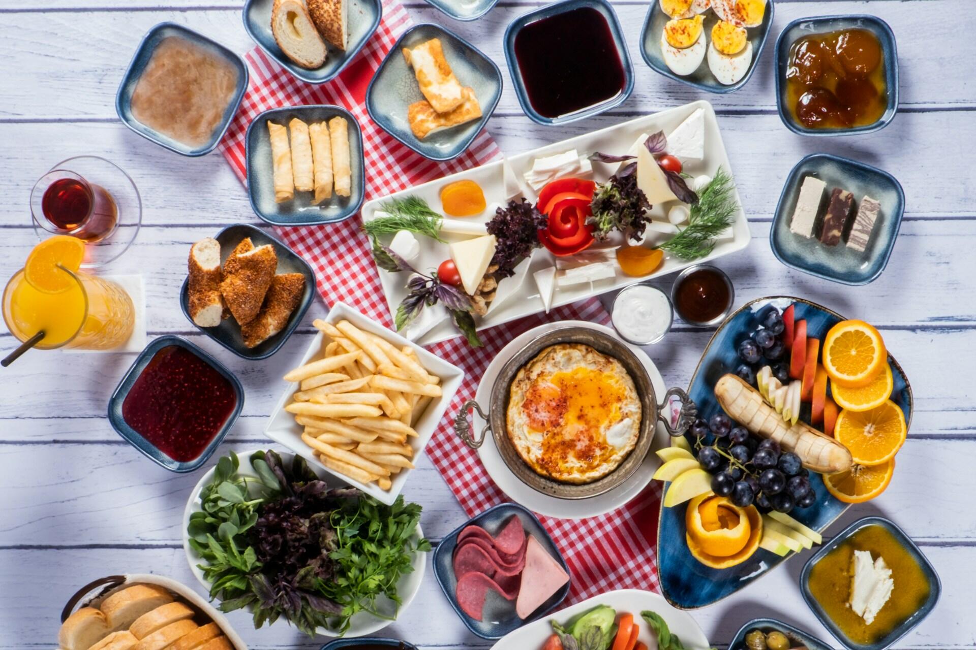 Bir sofrada Türk kahvaltısına ait zengin çeşitler: peynirler, zeytinler, yeşillikler, haşlanmış yumurta, sahanda yumurta, börek, reçeller, bal, taze meyveler, kızarmış patates, simit ve çay eşliğinde renkli bir sunum.