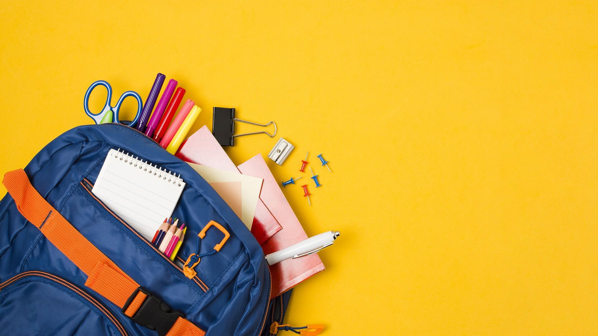 Mavi okul çantasının içinden çıkan defter, kalem, makas ve kırtasiye malzemeleri, sarı arka plan üzerinde.
