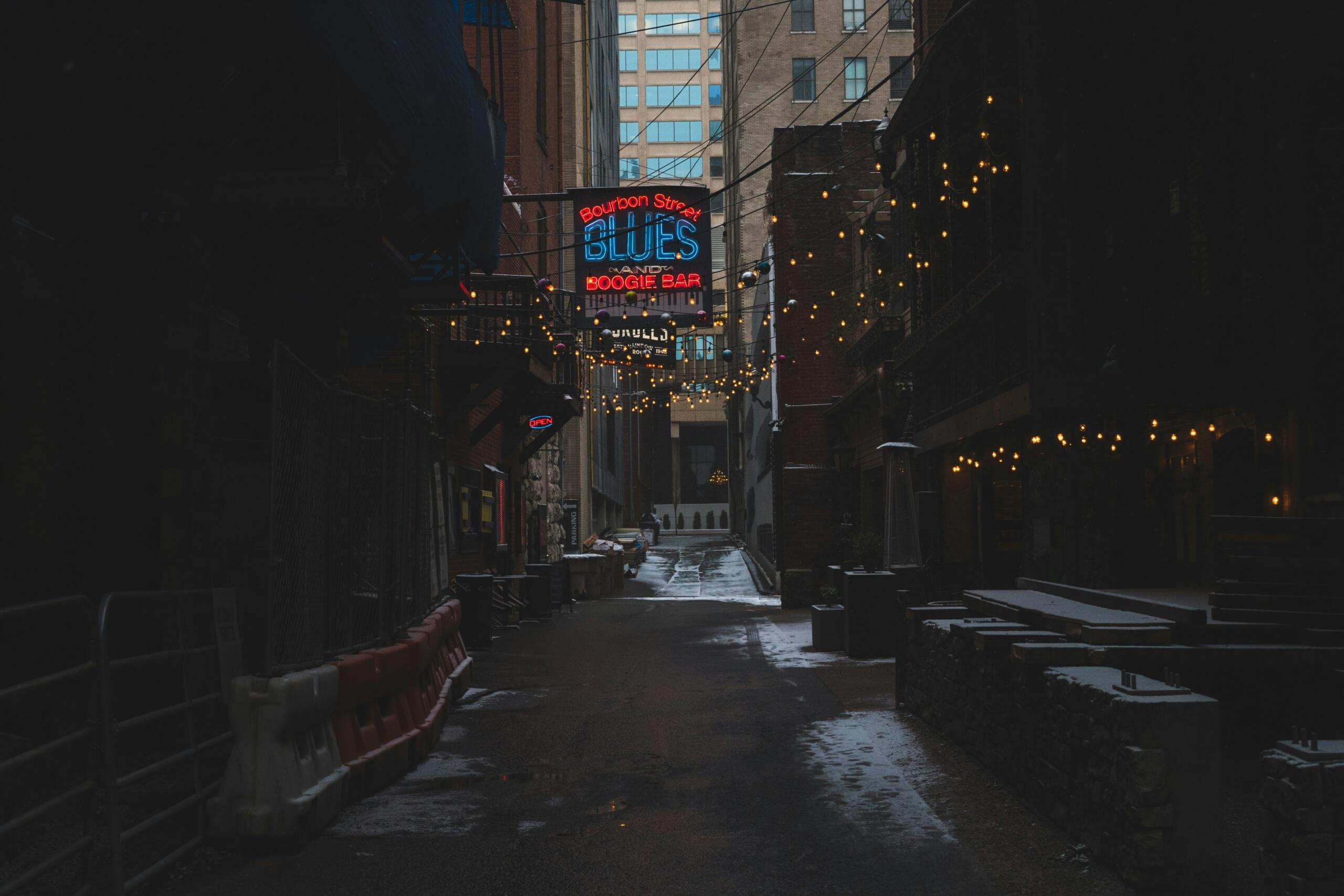 Dar bir sokakta asılı ışıklarla süslenmiş bir geçit; ortada 'Bourbon Street Blues and Boogie Bar' yazılı neon tabela parlıyor.