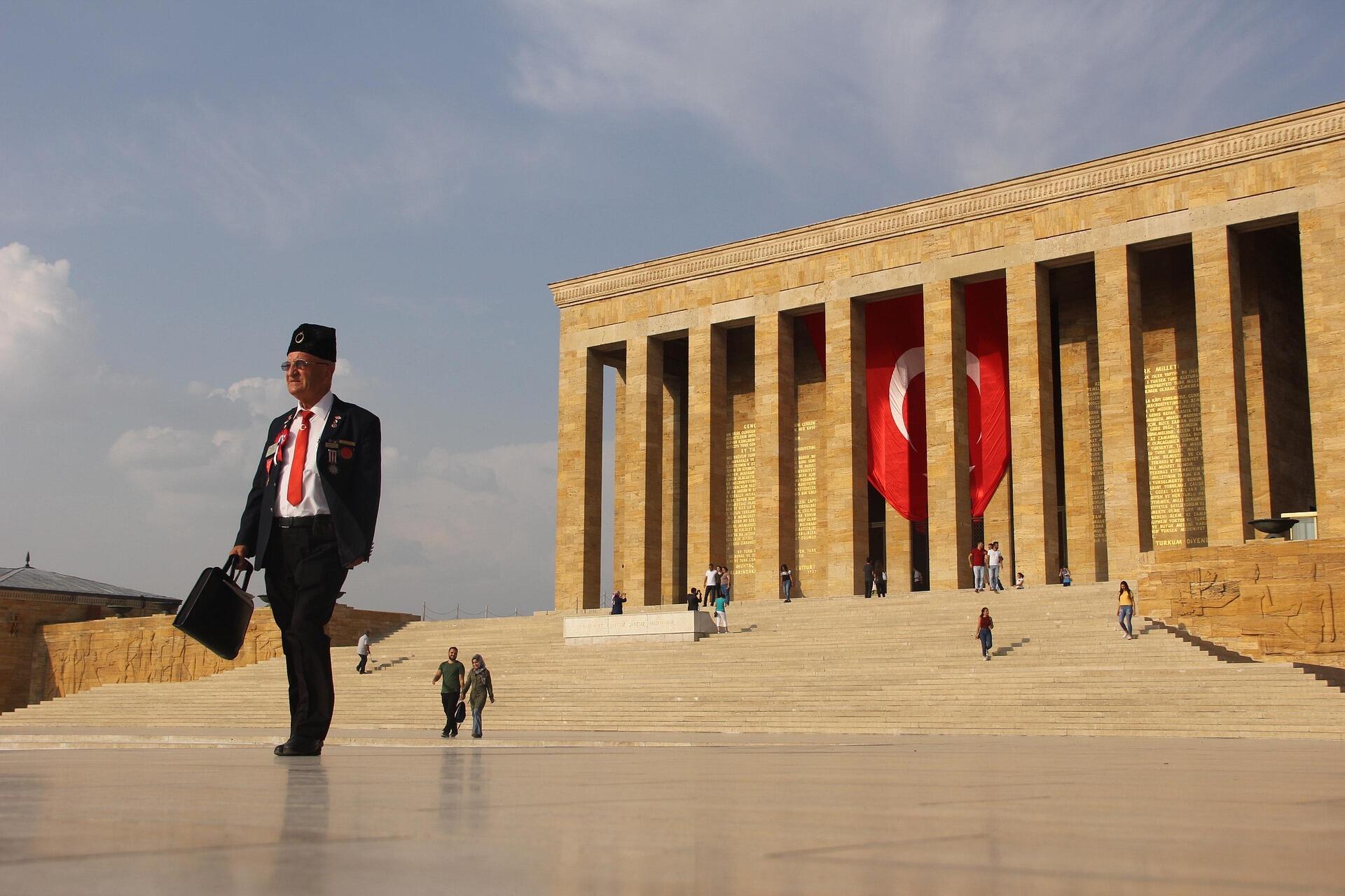 Bir Türk gazisi, elinde çantasıyla Anıtkabir’in önünden geçerken; arka planda dev Türk bayrağı ve mozoleyi ziyaret eden insanlar görülüyor.