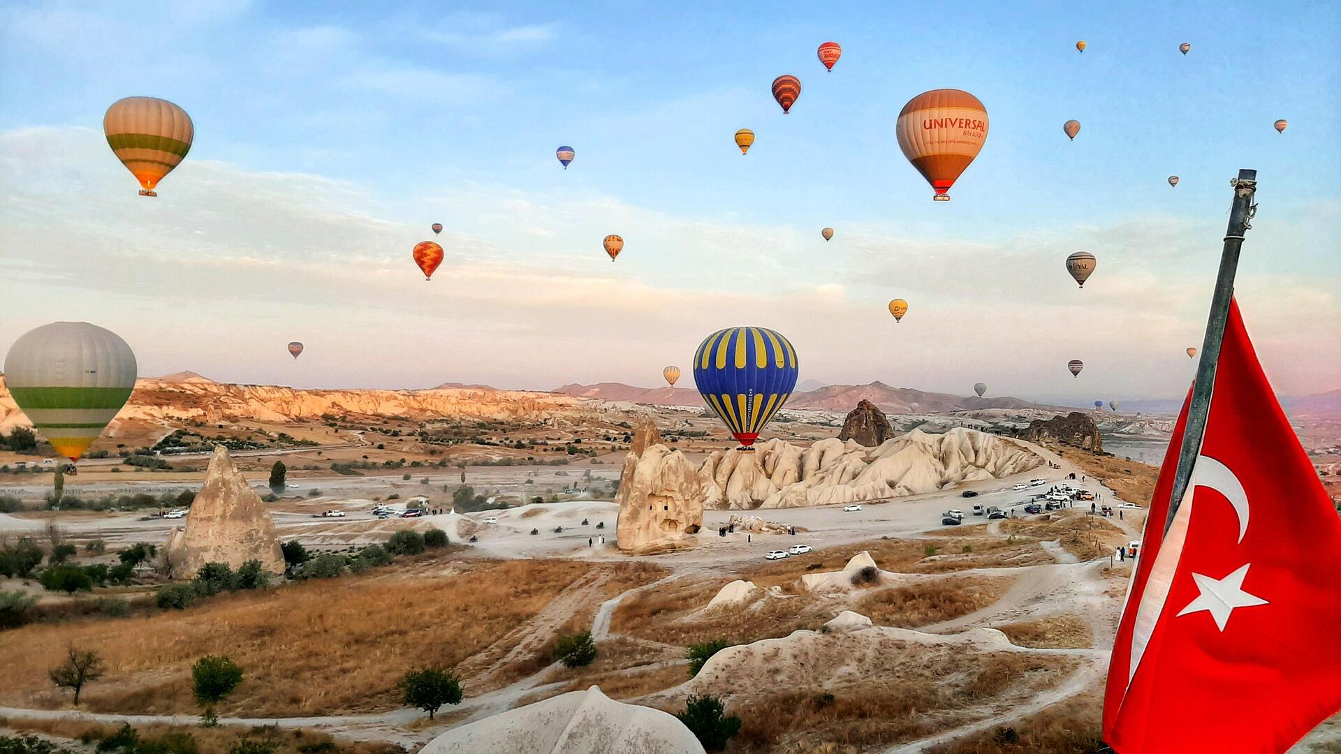 Kapadokya’da gün doğumunda gökyüzünde süzülen rengarenk sıcak hava balonları ve ön planda dalgalanan Türk bayrağı.