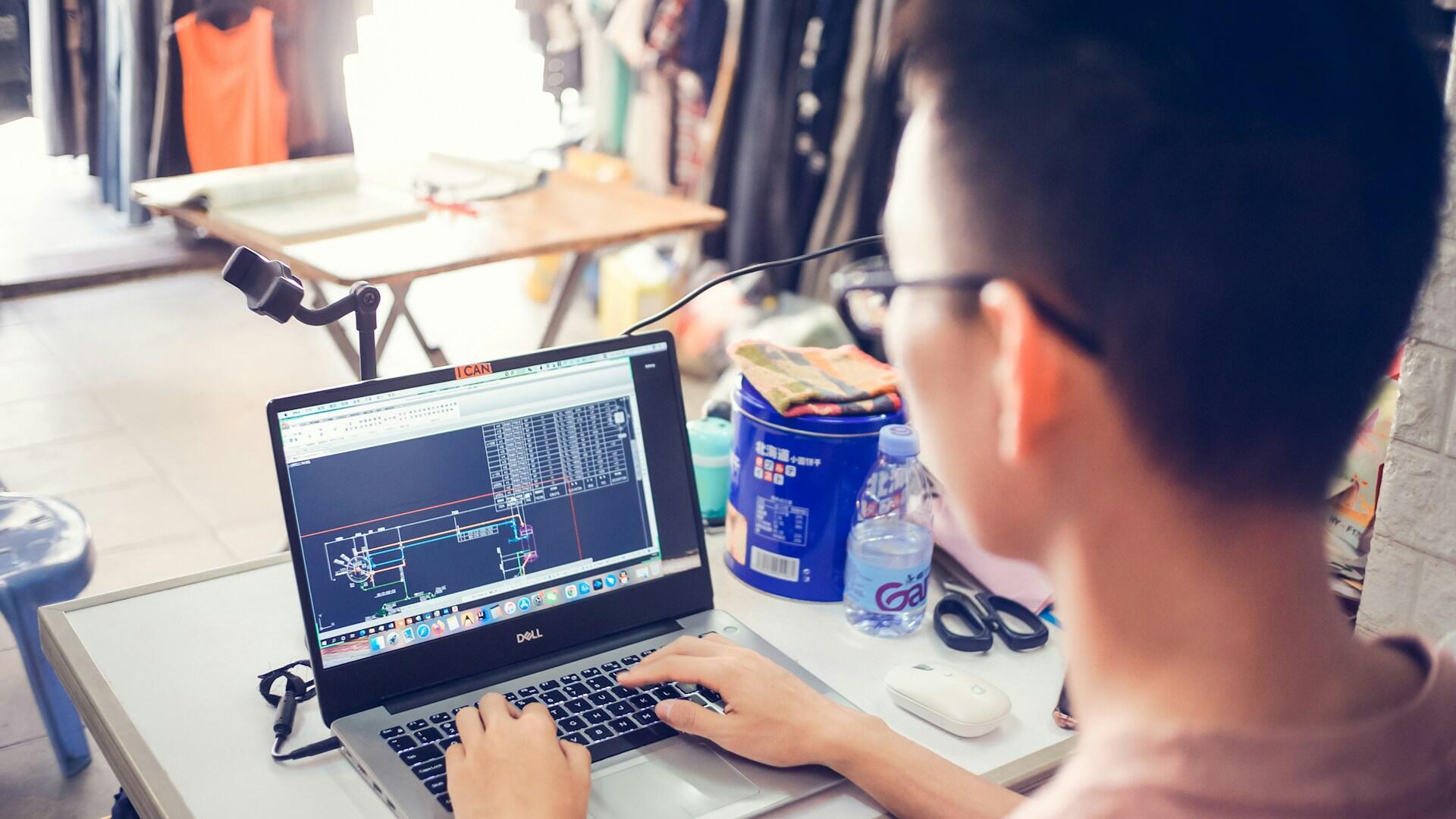 "Bir masada arkadan görünen genç bir adam, laptop ekranındaki siyah zeminli AutoCAD teknik çizim programına odaklanmış, klavyeyi kullanıyor. Dağınık bir çalışma ortamı.