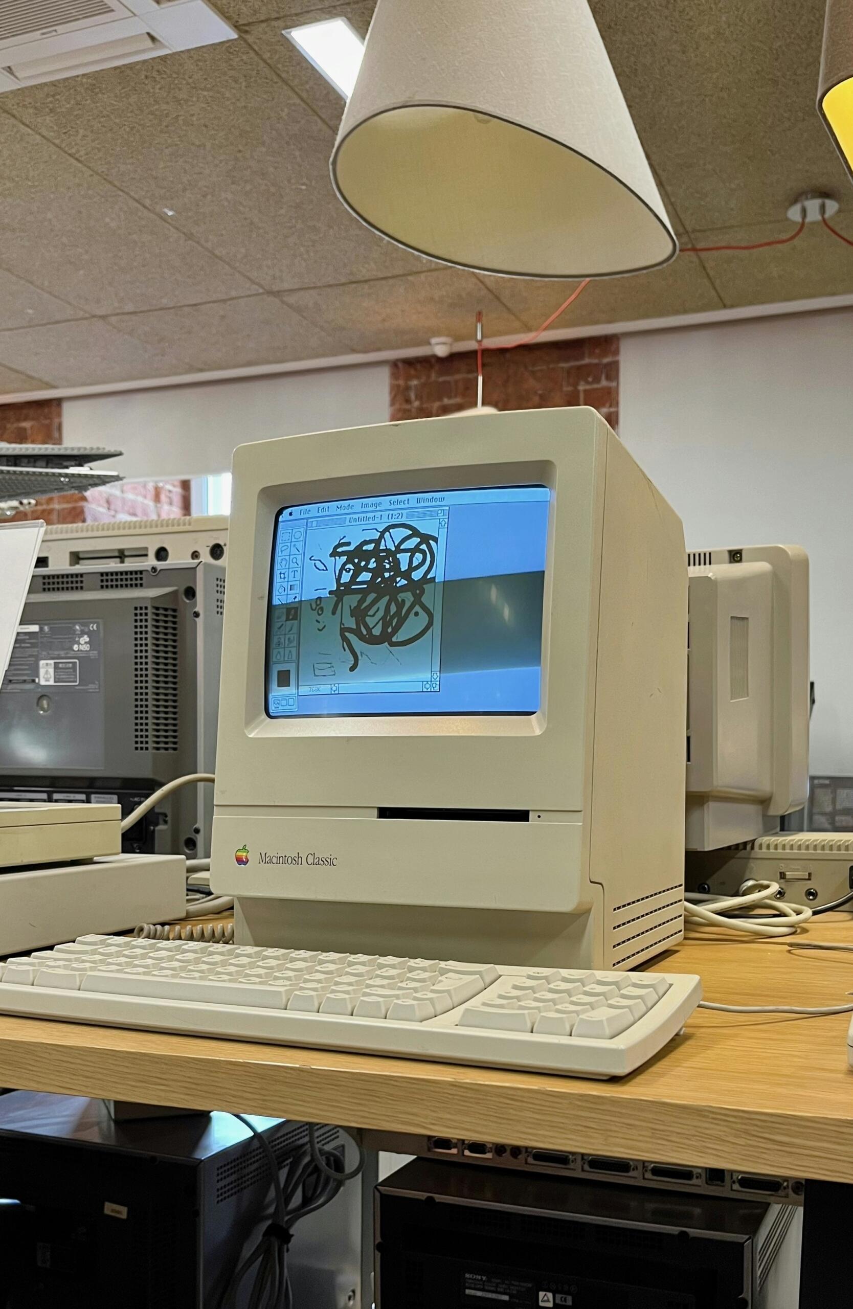 Bir lamba altında, bir masanın üzerinde duran, ekranında karalama yapılmış eski bir Macintosh Classic bilgisayar ve klavye.
