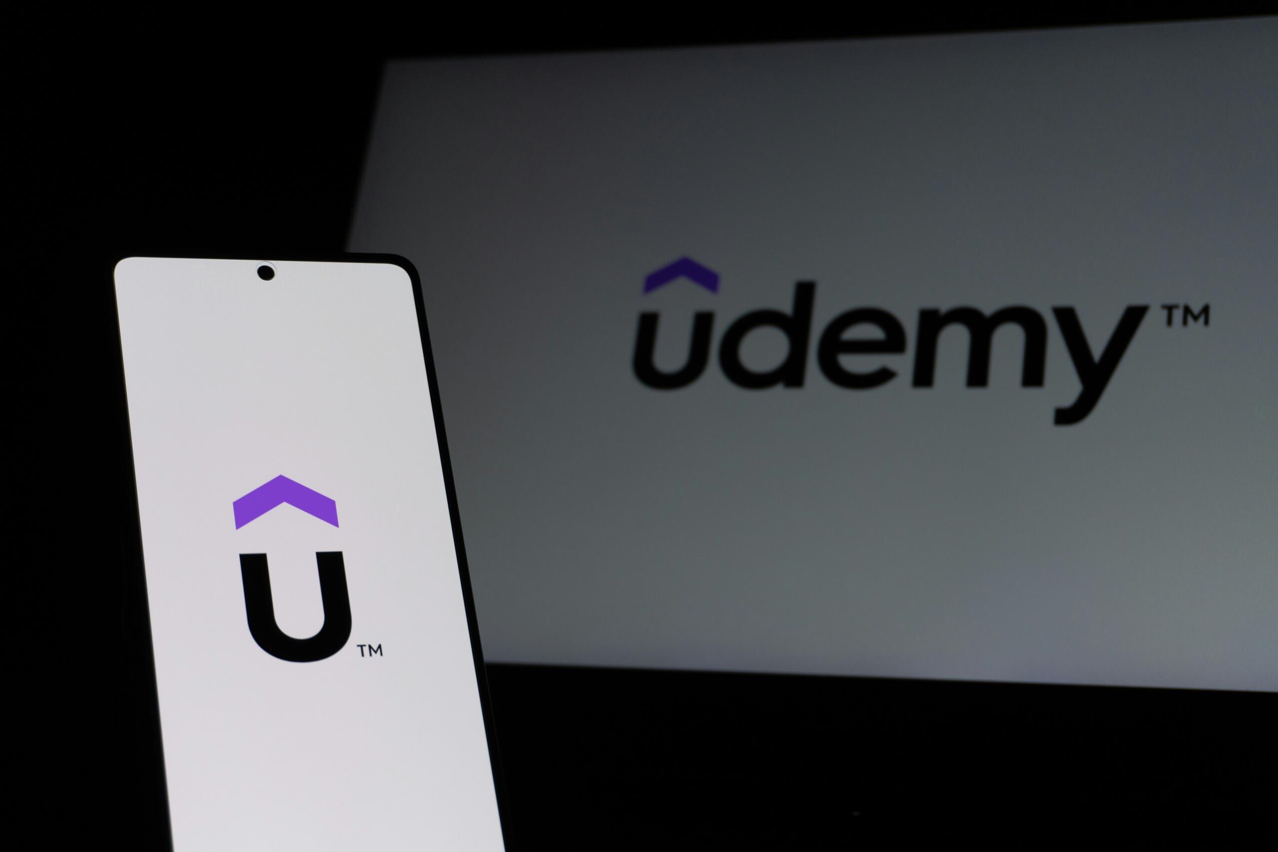 Görsel, ön planda Udemy mobil uygulamasının logosu ve adının bulunduğu bir akıllı telefonu, arka planda ise aynı platformun bilgisayar ekranındaki logosunu ve adını gösteriyor.