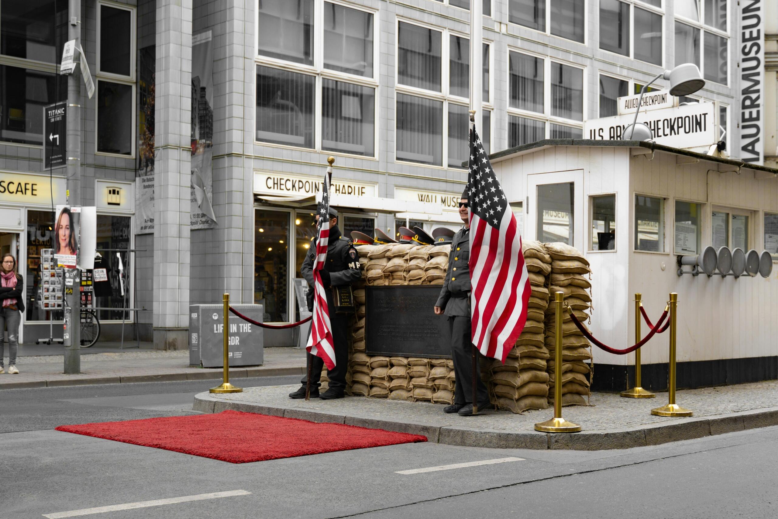 Berlin’deki Checkpoint Charlie’de, Amerikan bayrakları taşıyan üniformalı görevliler ve eski sınır kulübesi görülüyor.