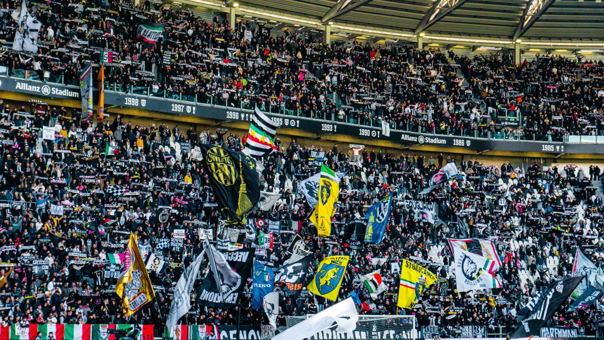 Juventus'un Allianz Stadium'unda Serie A maçı atmosferi. Tribünler siyah-beyaz atkılar, Juventus bayrakları ve renkli koreografiyle dolup taşıyor. Duvarlarında 1985'ten 1998'e uzanan şampiyonluk yılları kazınmış olan bu stat, İtalya ligi Serie A'nın en ikonik sahnelerinden birini sunuyor.