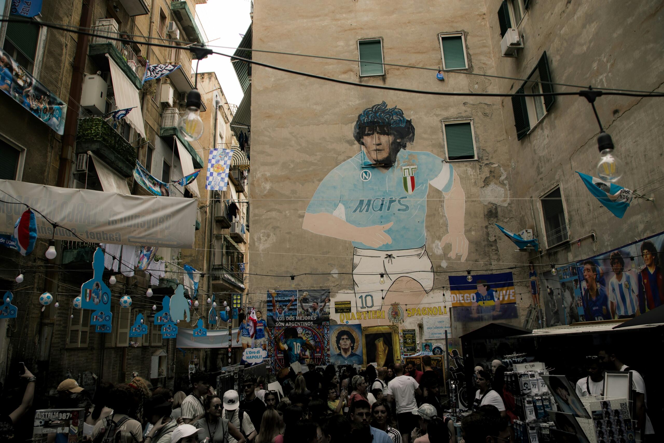 Napoli sokaklarında bir binanın duvarına yapılmış dev Maradona duvar resmi. Önünde İtalyan bayrakları, Napoli formaları ve hediyelik eşyalar satılan canlı bir pazar yeri. Fotoğraf, İtalya ligi Serie A'nın sadece bir spor değil, bir kültür ve yaşam biçimi olduğunu gözler önüne seriyor.