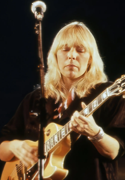 Ünlü sanatçı Joni Mitchell sahnede gitar çalarken; samimi ve duygusal bir performans anını yansıtan nostaljik sahne fotoğrafı.