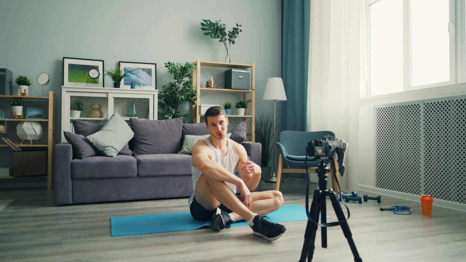 Oturma odasındaki yoga matı üzerinde, kameraya (tripodlu) bakarak konuşan ve antrenman anlatan erkek online fitness koçu.