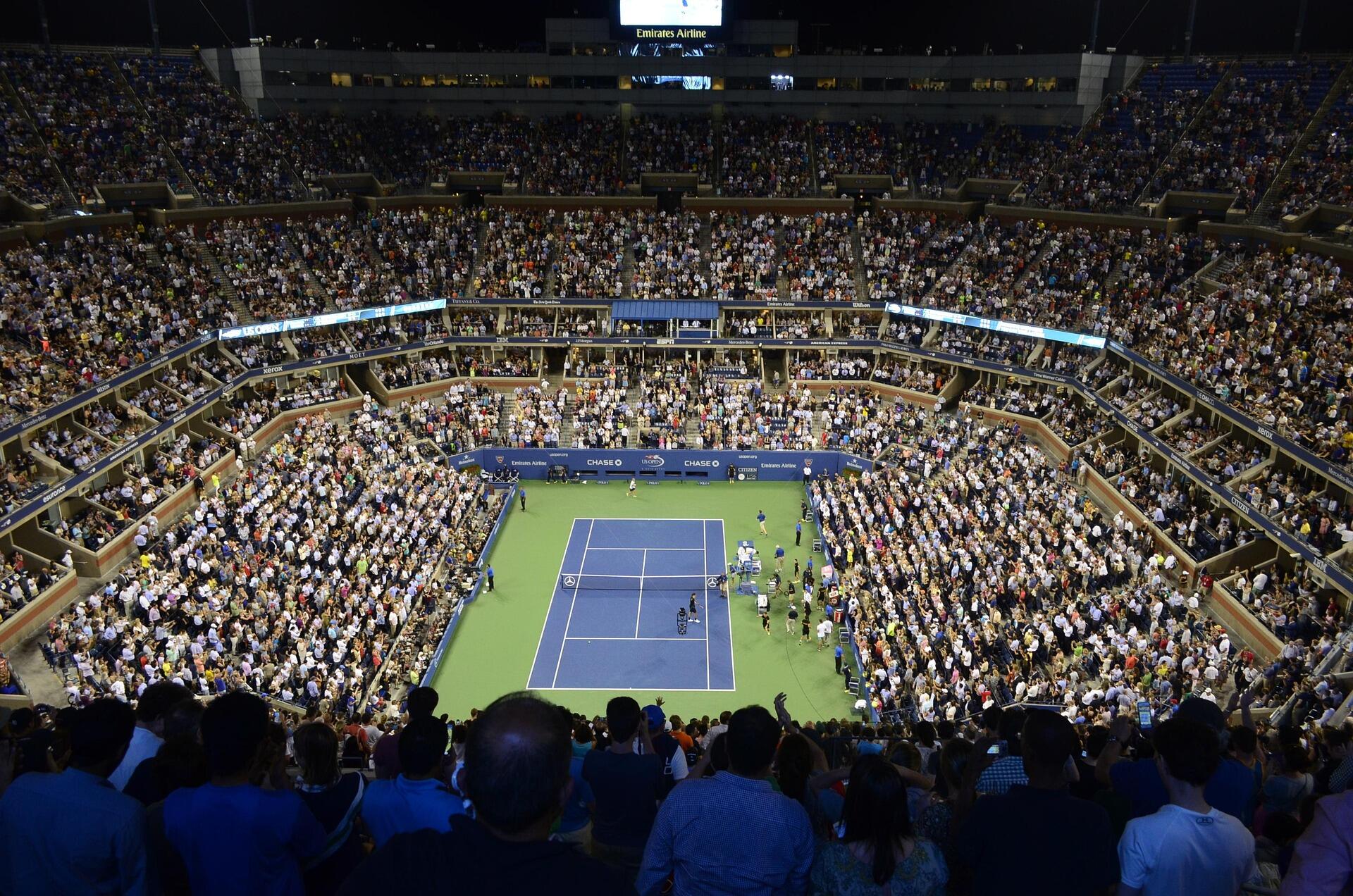 Tıklım tıklım dolu tribünlerle çevrili, gece ışıklandırılmış bir Grand Slam tenis kortu stadyumu.