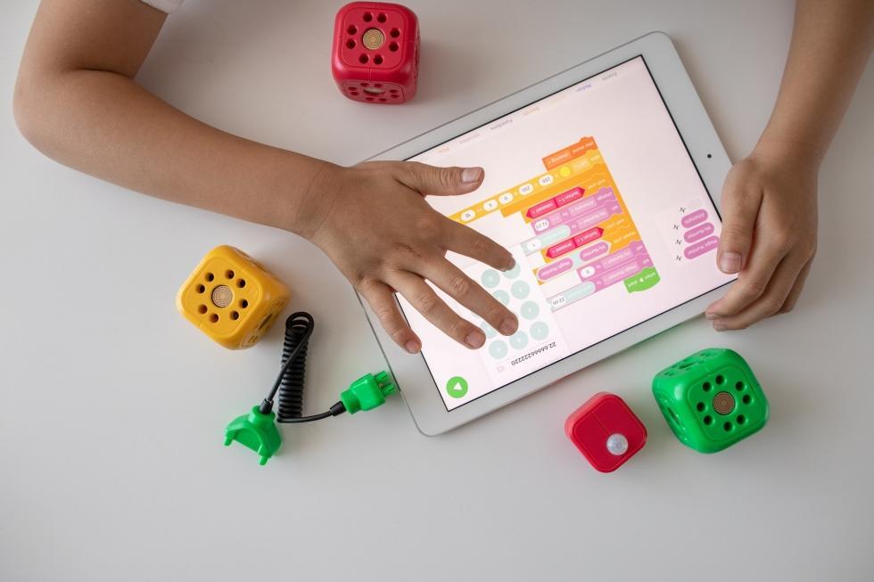Bir çocuğun tablet üzerinde renkli bir matematik/öğrenme uygulamasını kullandığı, etrafında sarı, kırmızı ve yeşil eğitim küplerinin bulunduğu masa üstü görüntüsü.