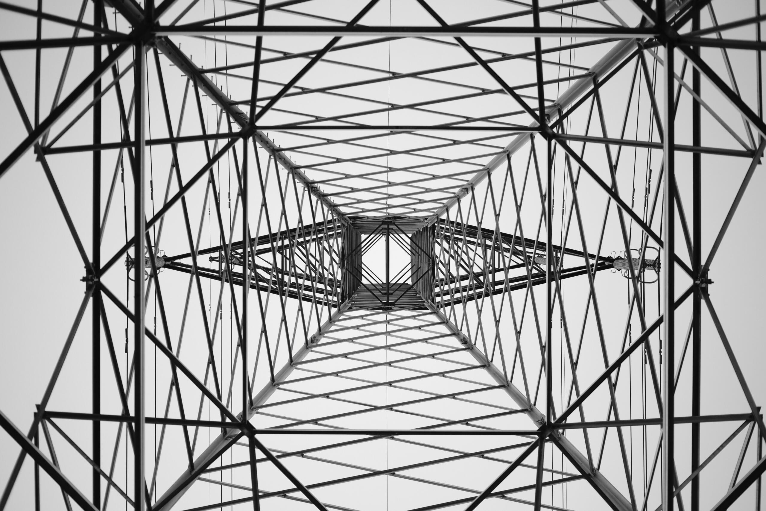 Alttan yukarı doğru çekilmiş, çelik bir elektrik iletim direğinin iç yapısını gösteren siyah-beyaz fotoğraf. Metal kirişler ve çapraz destekler merkezde geometrik, simetrik bir perspektif oluşturuyor.