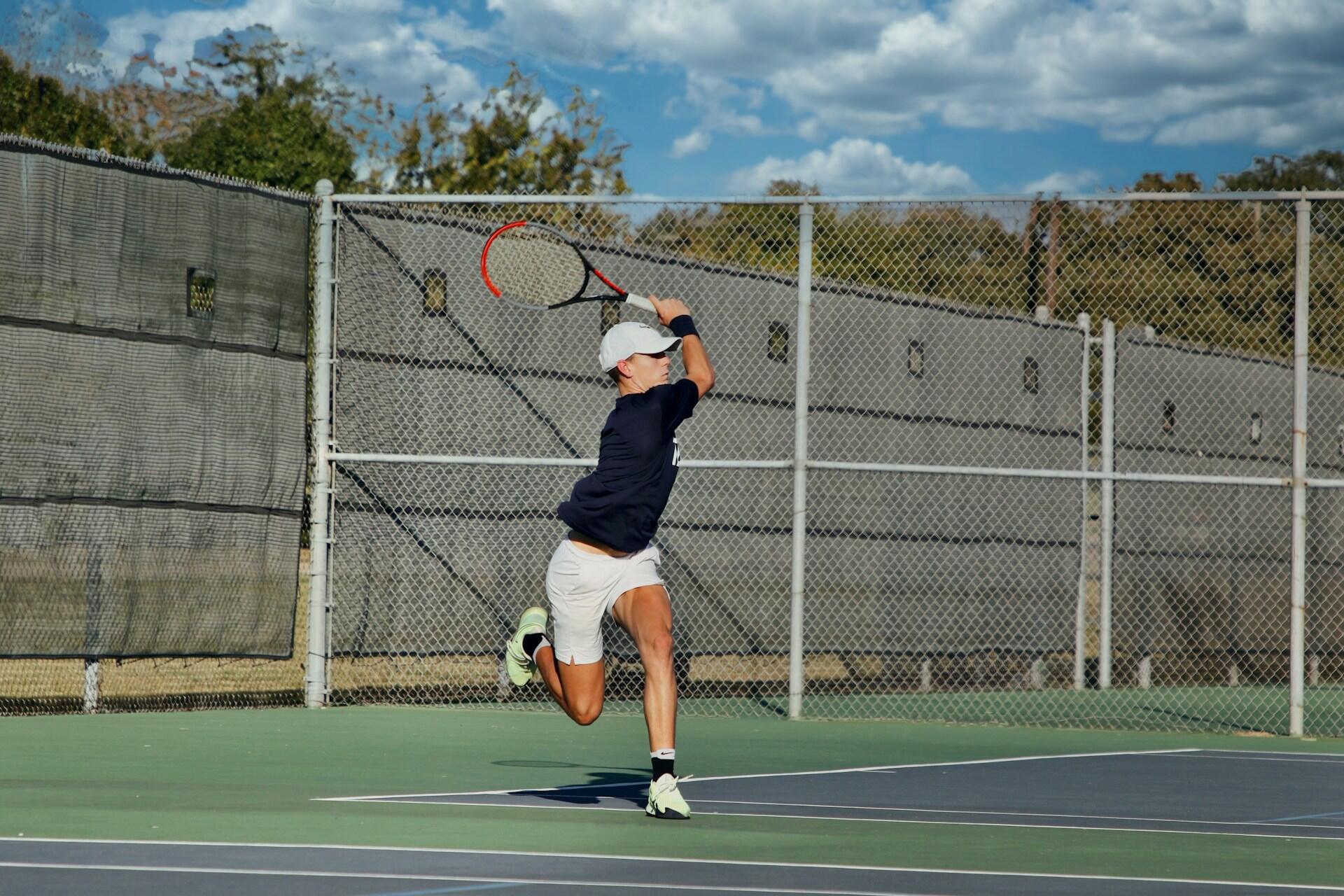 Açık tenis kortunda, siyah tişört, beyaz şort ve şapka giyen bir erkek tenisçinin, arka el (backhand) vuruşu yapmak için havada sıçradığı anın dinamik fotoğrafı. Arka planda tel örgü, yeşil branda, ağaçlar ve parçalı bulutlu mavi gökyüzü görülüyor.