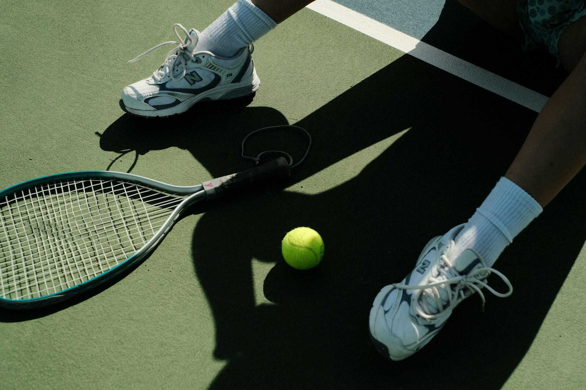 Yeşil renkli sert tenis kortu zemininde oturan bir sporcunun bacakları, beyaz spor ayakkabıları ve beyaz çorapları; hemen yanlarında yerde duran turkuaz çerçeveli bir tenis raketi ve parlak sarı bir tenis topunun yukarıdan çekilmiş yakın plan fotoğrafı.