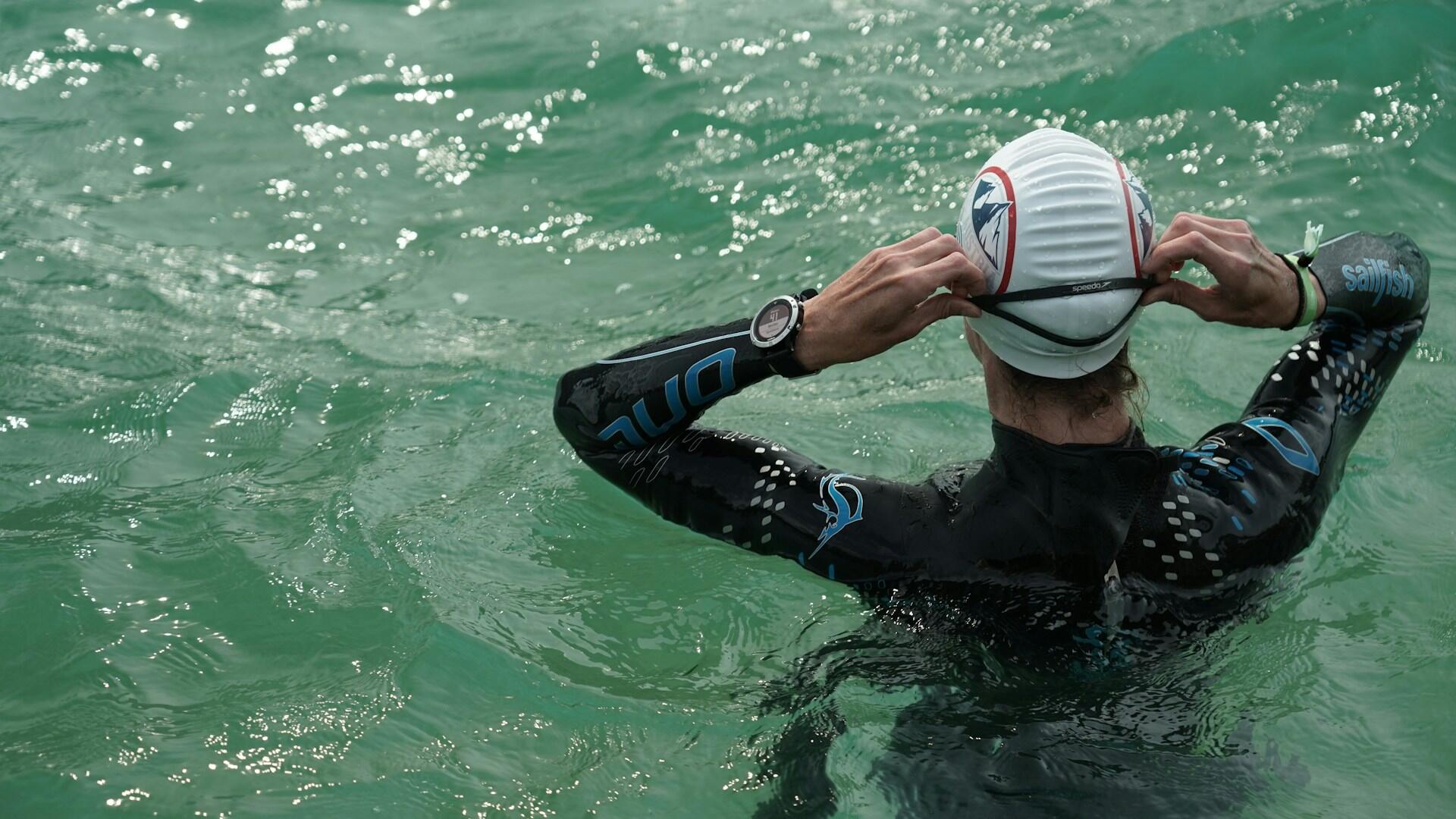 Açık deniz yüzme yarışına hazırlanan, yüzücü saati ve tam wetsuit giymiş bir yüzme antrenörü veya sporcusu, yeşilimsi dalgalı suyun içinde bonesini ve gözlüğünü takarken arkadan görünüyor.