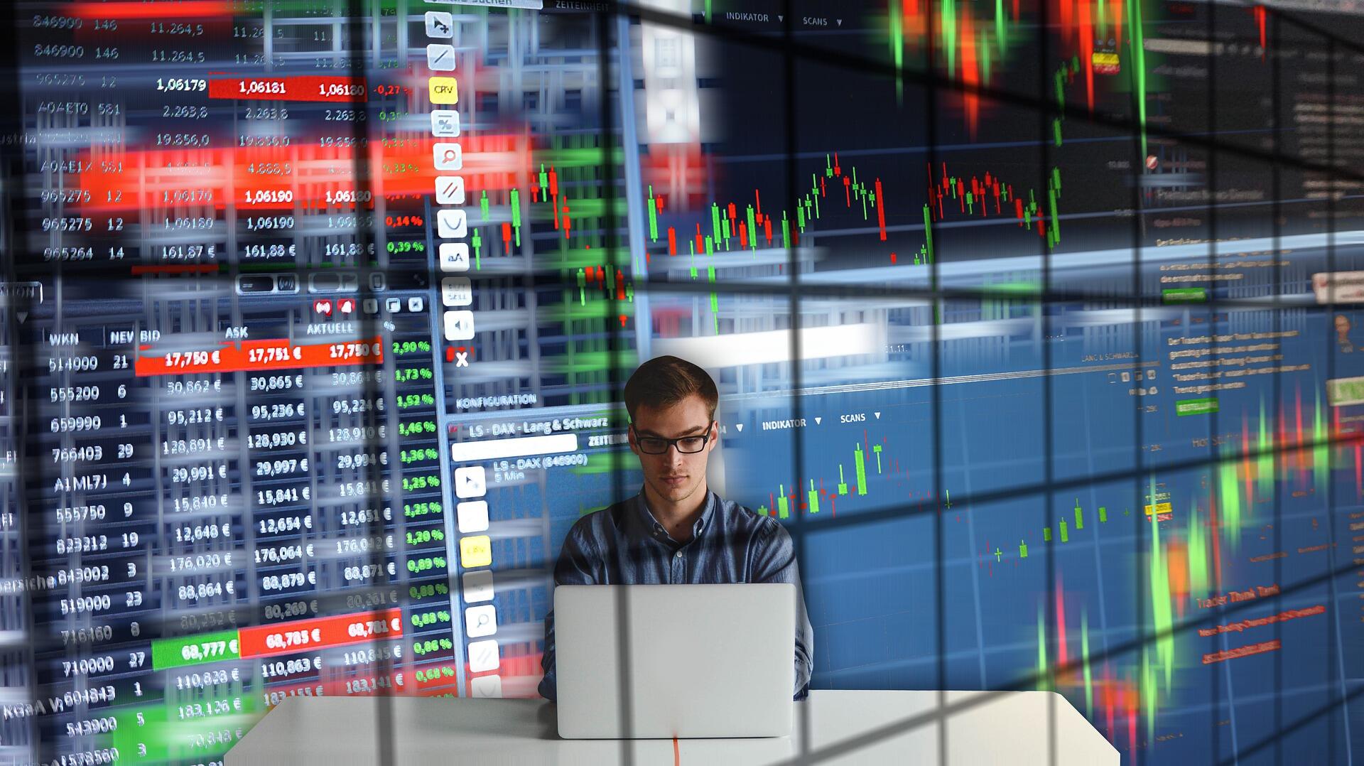 Dizüstü bilgisayar kullanan genç bir adamın, borsa verileri, hisse senedi fiyatları ve finansal grafiklerin canlı olarak yansıtıldığı büyük bir veri ekranının önünde oturduğu kompozit bir görüntü.