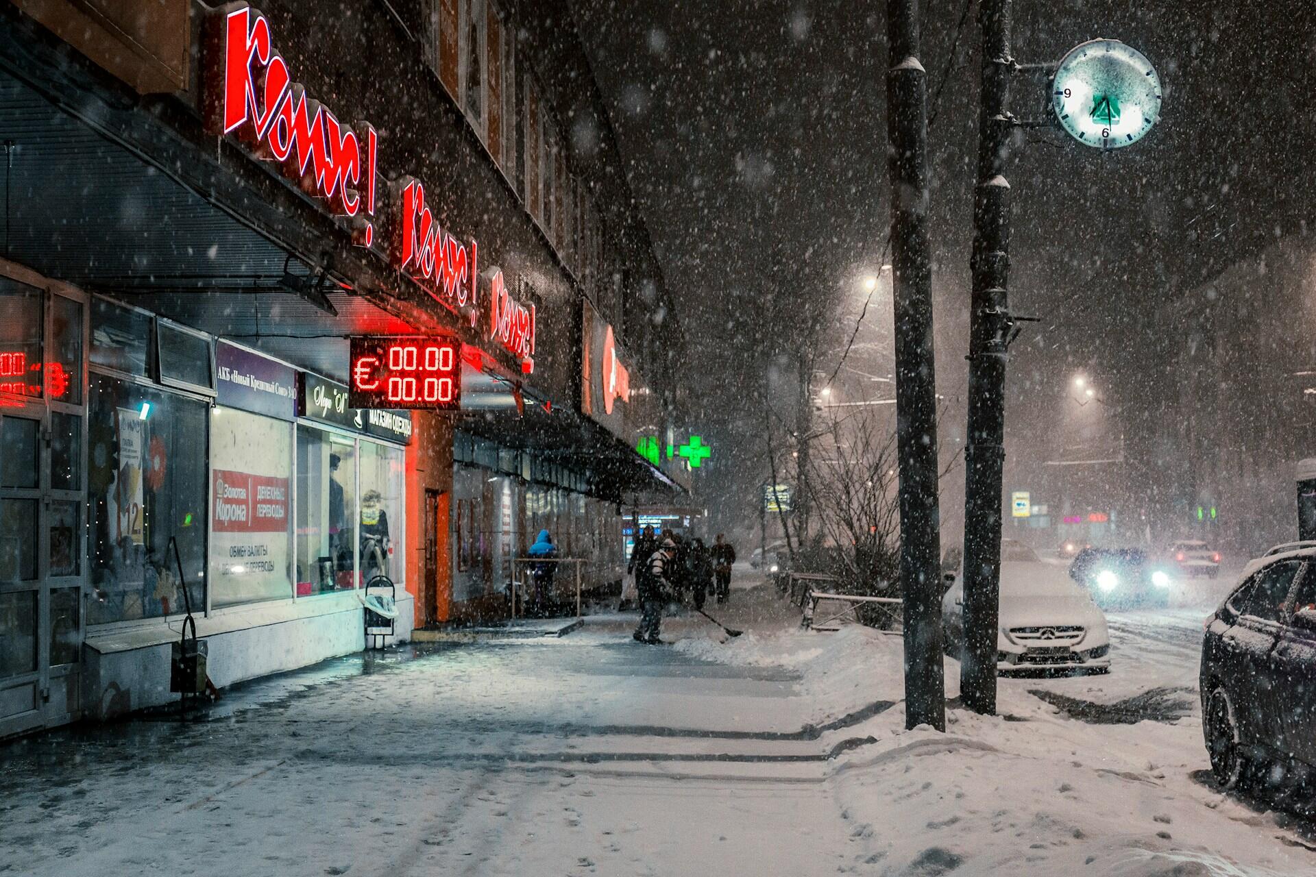 Yoğun kar yağışı altında gece vakti bir Rus şehri caddesi. Solda üzerinde Kiril alfabesiyle kırmızı neon tabelalar ve döviz kurları bulunan mağazalar, kaldırımda kar küreyen bir kişi ve caddede ışıkları yanan arabalar görülüyor.