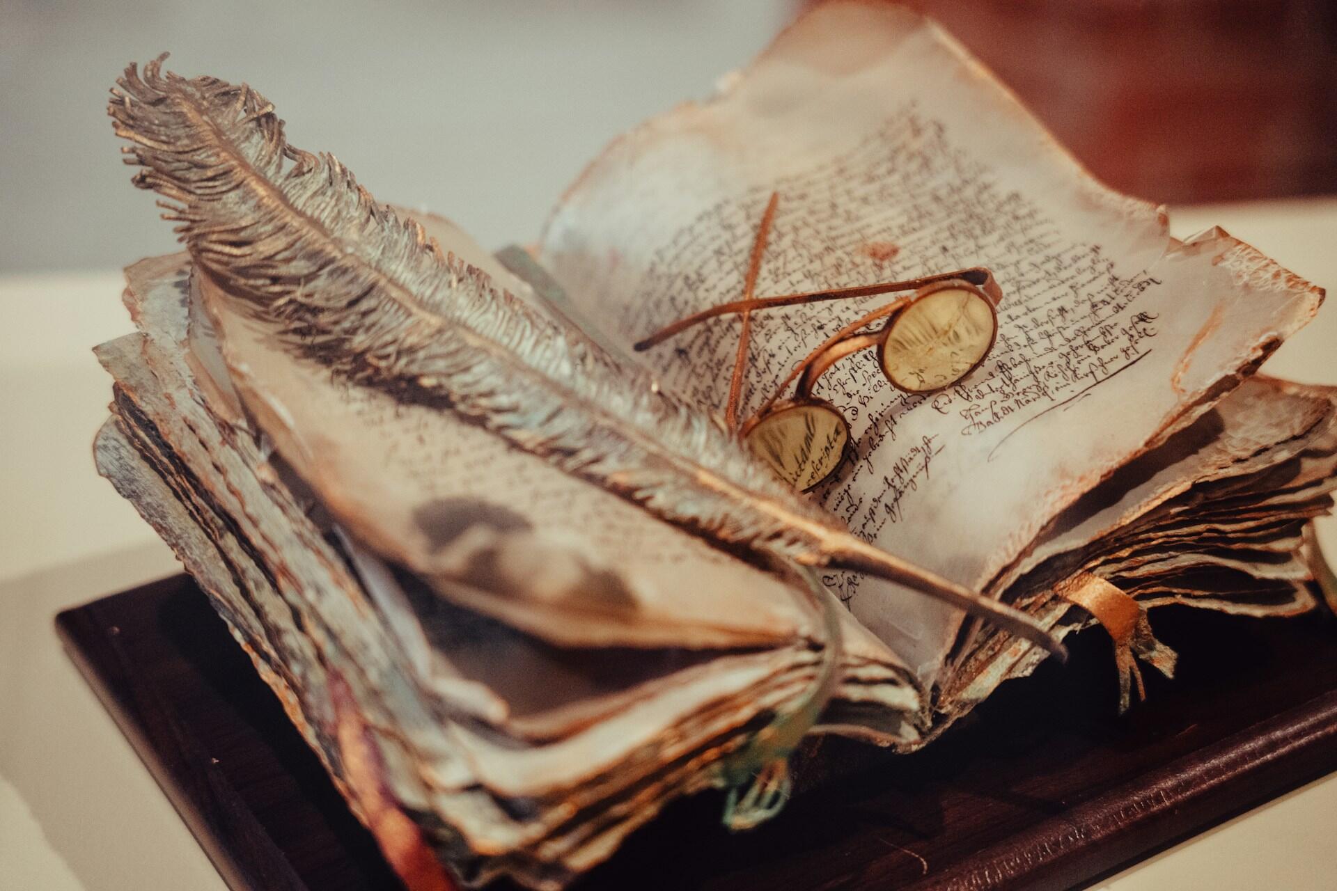 розгорнута антикварна книга у шкіряній обкладинці, між сторінок лежать перо для письма та вінтажні окуляри