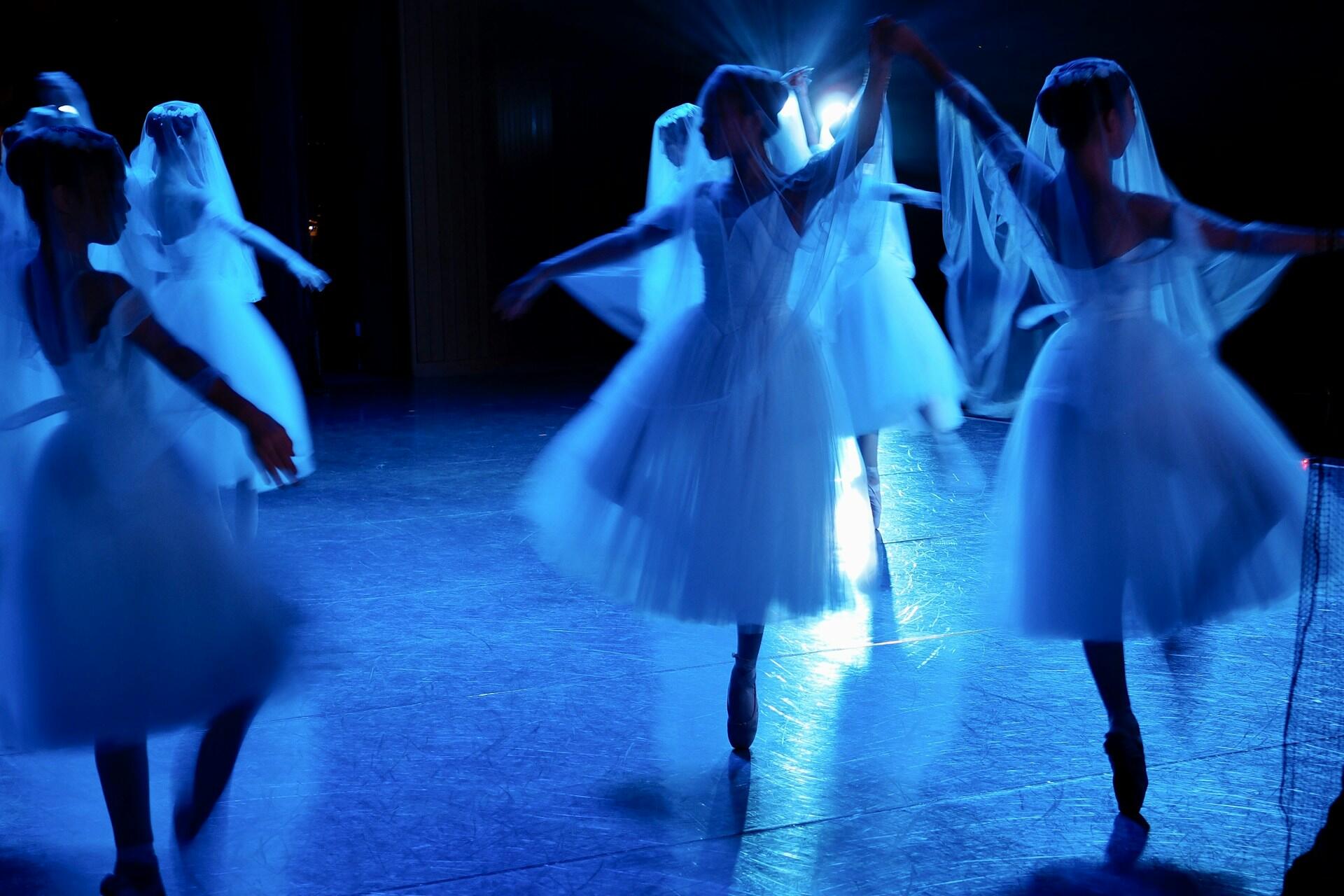 Ballerina's on stage with white costumes and a blue light shining on them.