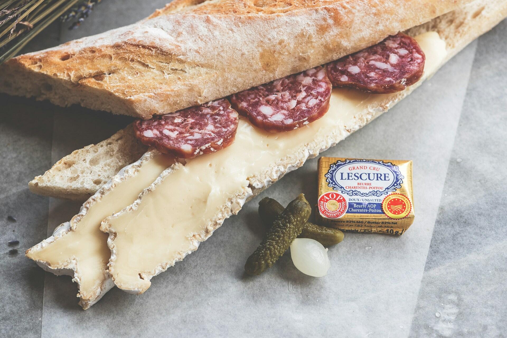 A baguette sandwich with brie and salami next to pickles and butter.