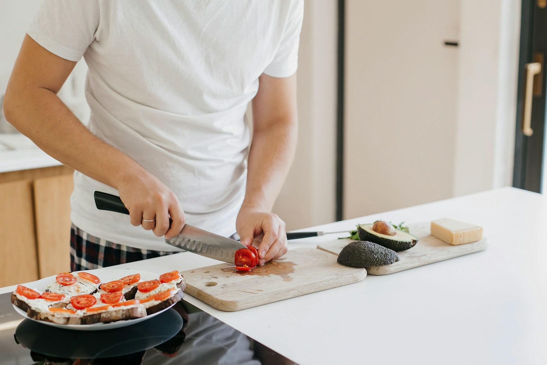 A man cutting up a tomato in the kitchen, with toast and avocado on the side.