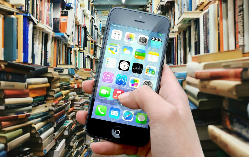 A hand is holding a cellphone in front of many books that surround it