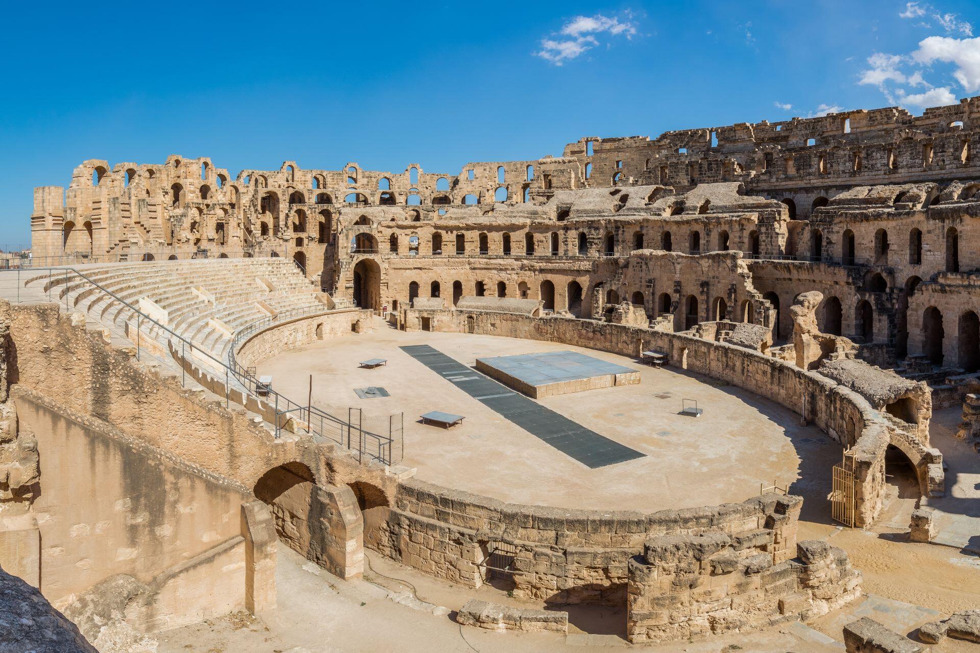 Wide view of a large Roman amphitheater with stone seating and arches under a clear blue sky.