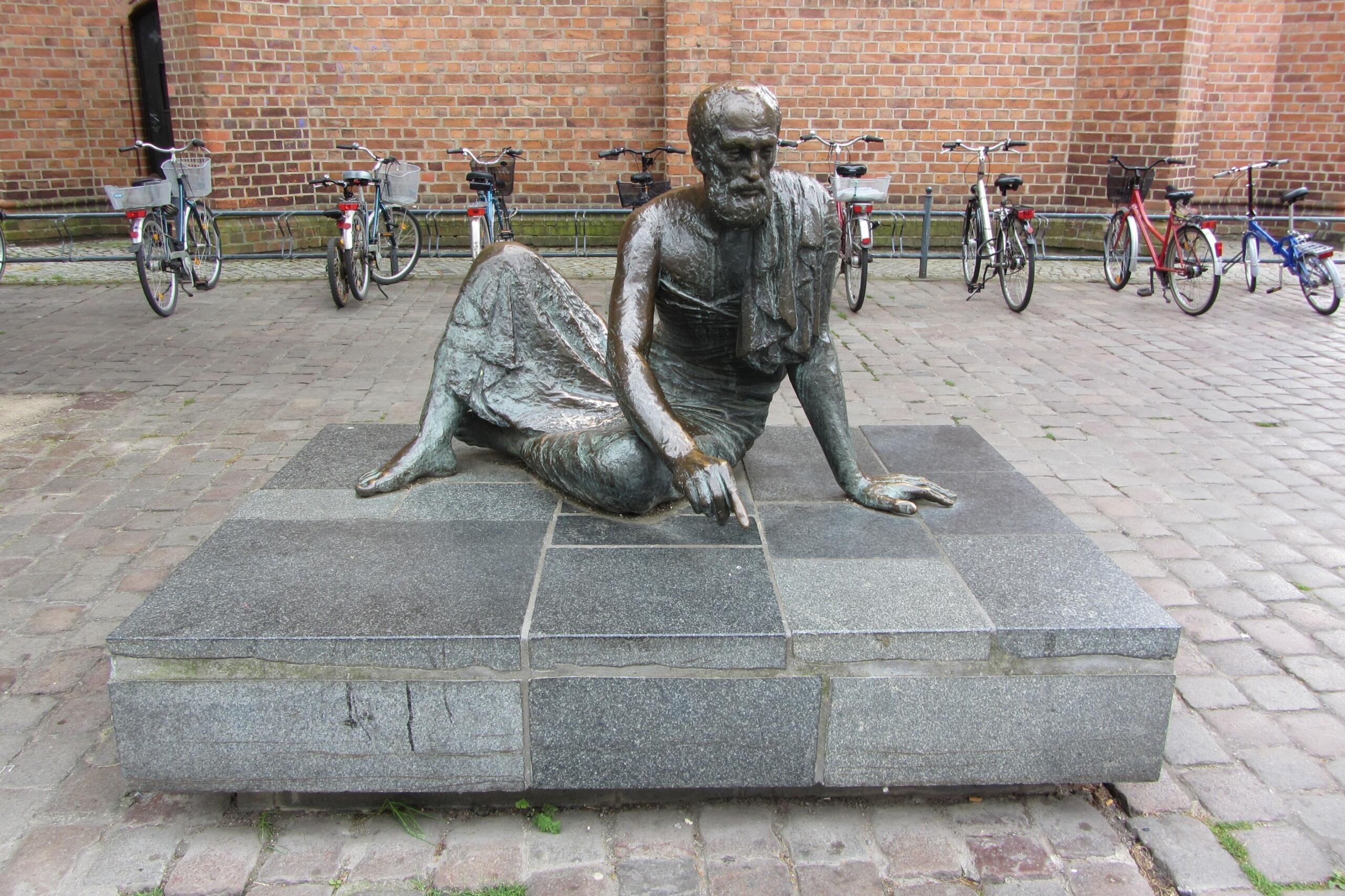 Bronze statue of Archimedes lying on the ground pointing at geometric shapes on a stone surface