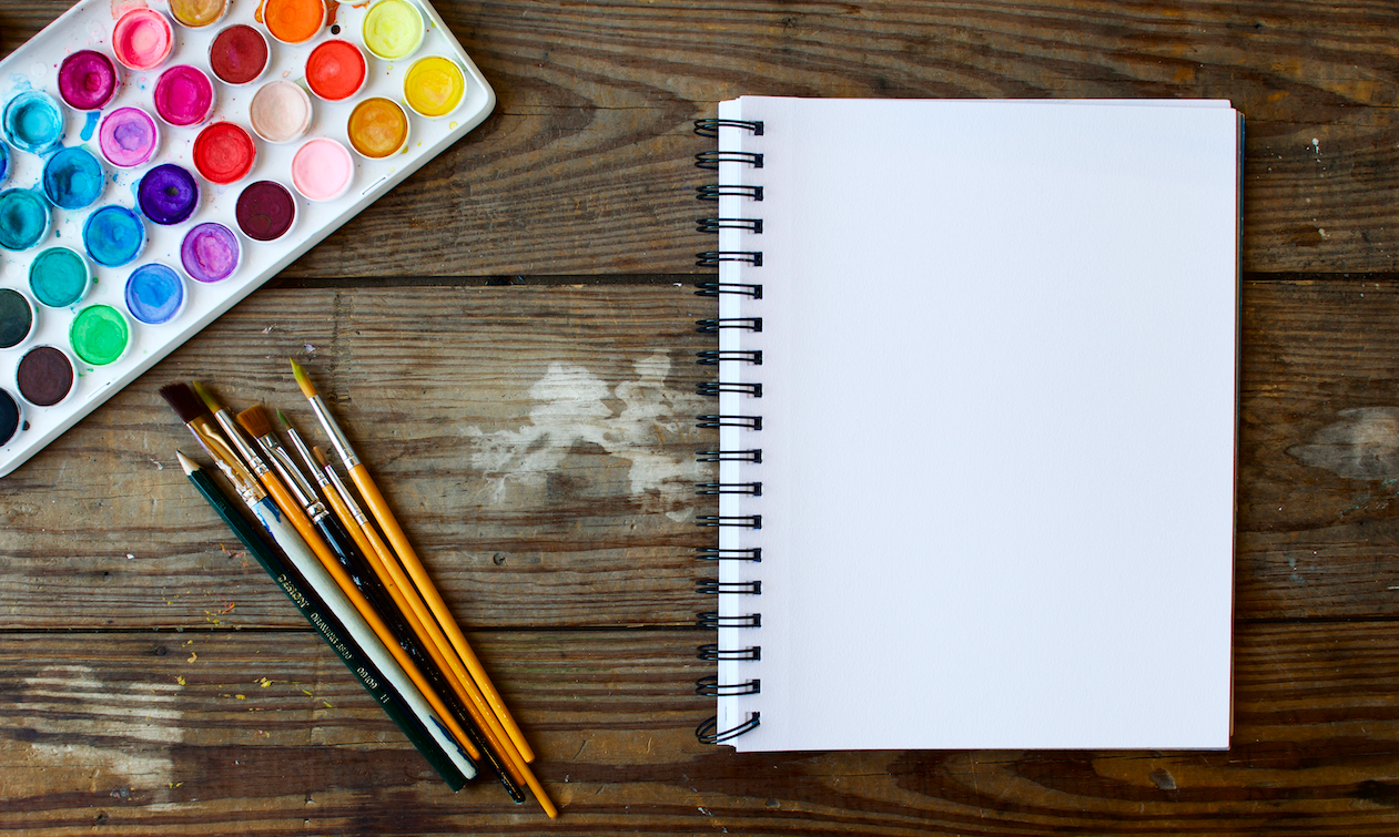 blank notebook page next to paintbrushes and watercolor palette on a wooden table surface.