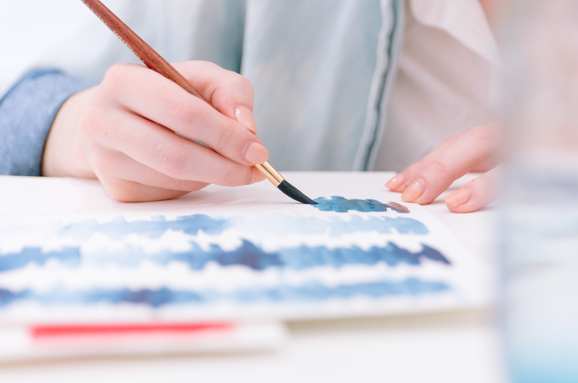 Person holding paintbrush and painting watercolor lines in blue.