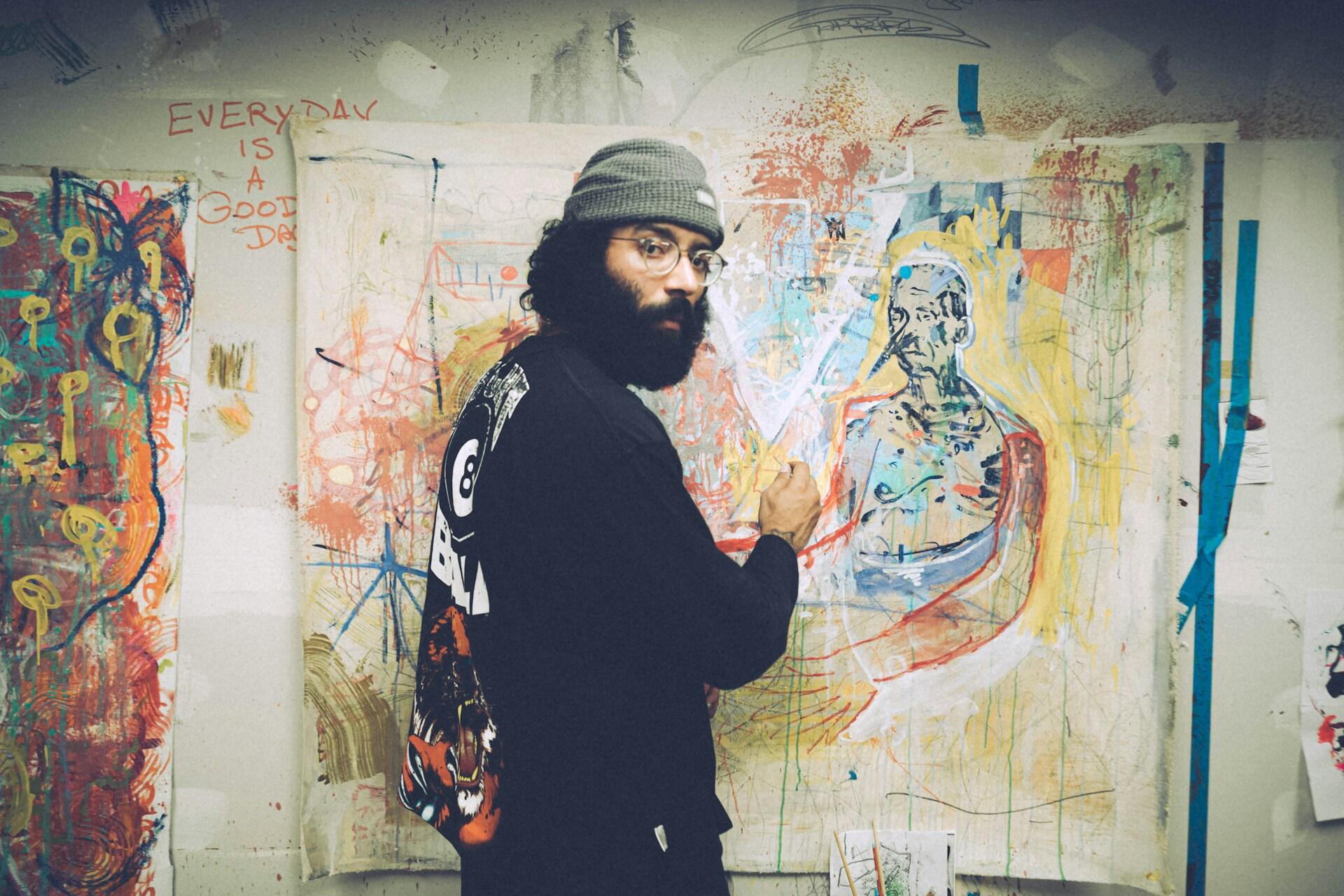 An artist in a beanie standing in front of his painting.