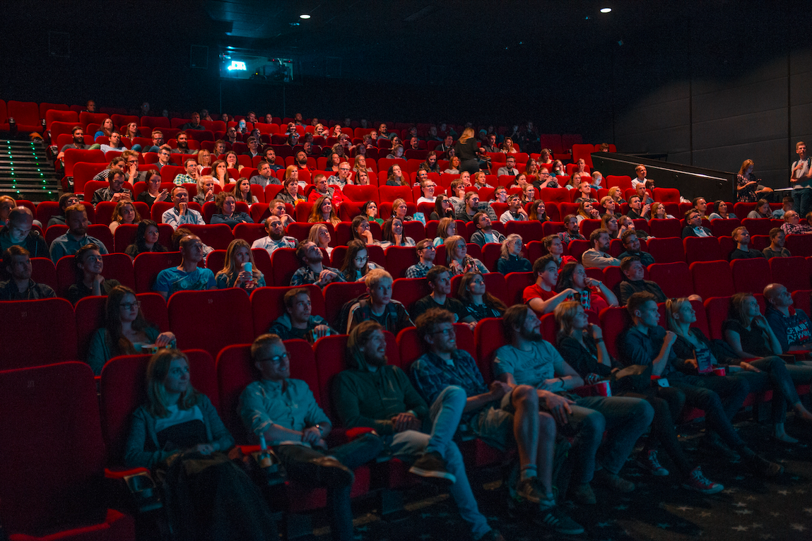 Many people sitting in the audience of a theatre on red chairs.